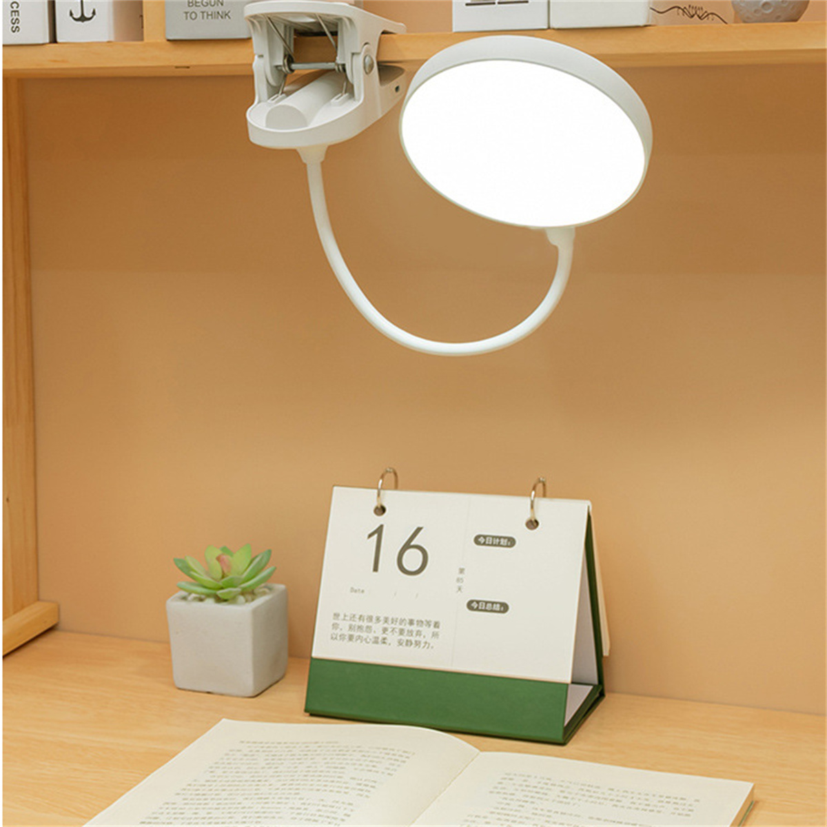 Eine weiße Schreibtischlampe mit Clip. Ein Kalender und eine kleine Pflanze stehen auf dem Schreibtisch. Der Hintergrund ist braun.