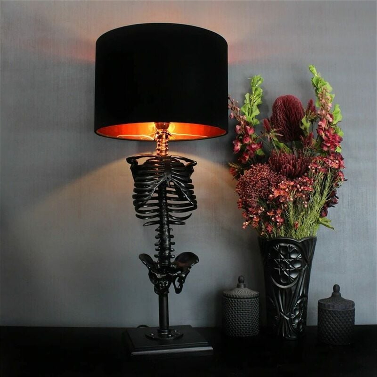 Skelettförmige Lampe mit schwarzem Lampenschirm. Blumen in einer Vase stehen neben der Lampe auf einem Tisch.