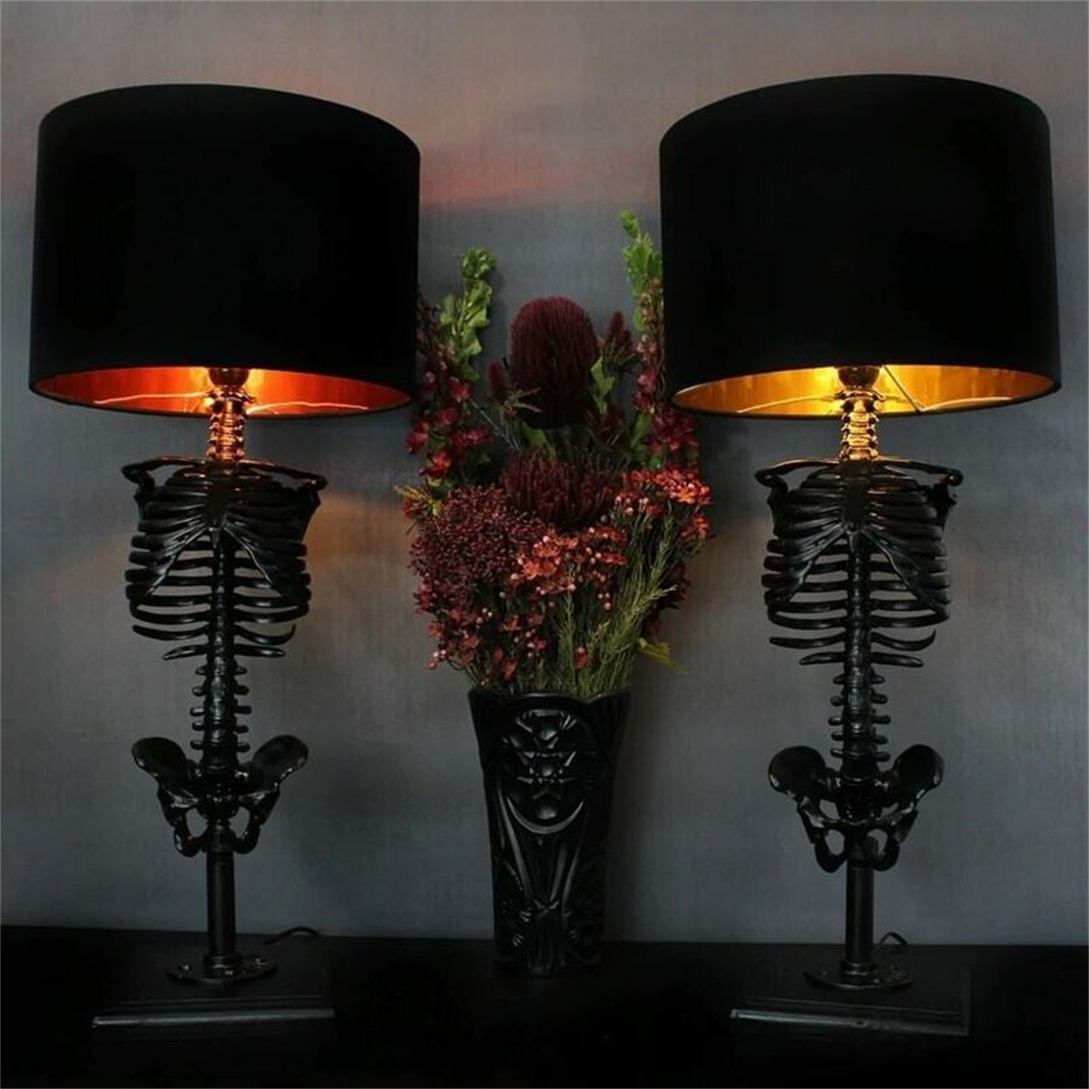 Zwei Skelettlampen mit schwarzen Schirmen und goldenem Innenraum flankieren eine Blumenvase auf einem Tisch.