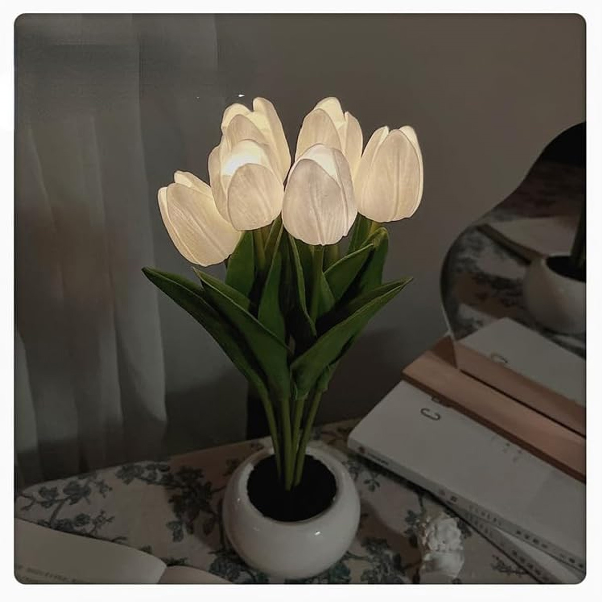 Ein Strauß weißer tulpenförmiger Lichter in einer weißen Vase auf einem Tisch mit Büchern.