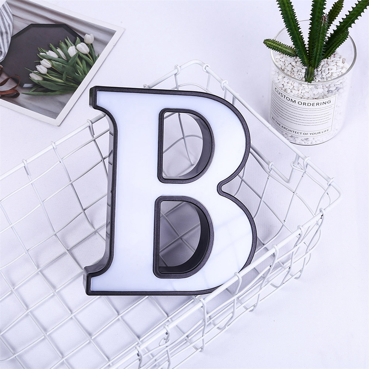 Weiß beleuchteter Buchstabe B in einem Drahtkorb mit weißen Blumen und einer Pflanze im Hintergrund.