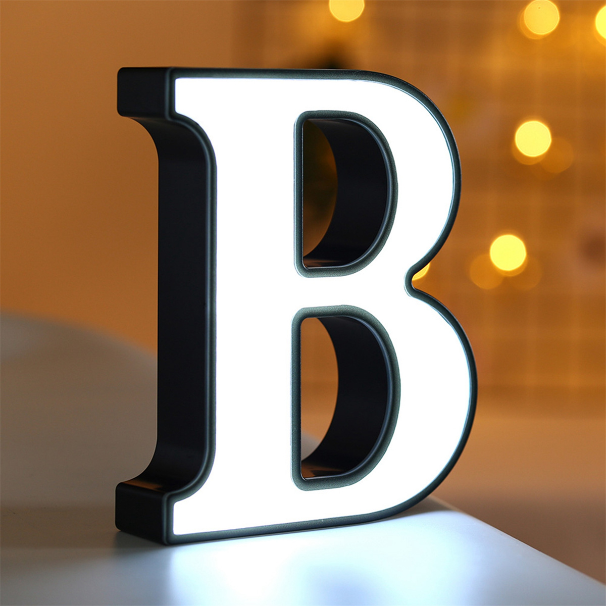 Ein weißer, beleuchteter Buchstabe B mit schwarzer Umrandung steht auf einem Tisch. Gelber Bokeh-Hintergrund.