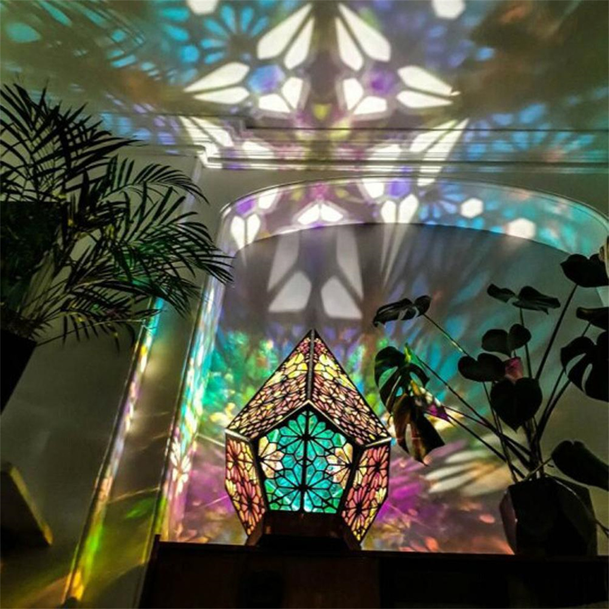 Eine geometrische Lampe mit farbenfroher Lichtanzeige, die Muster an die Wand wirft. Pflanzen befinden sich im Vordergrund.