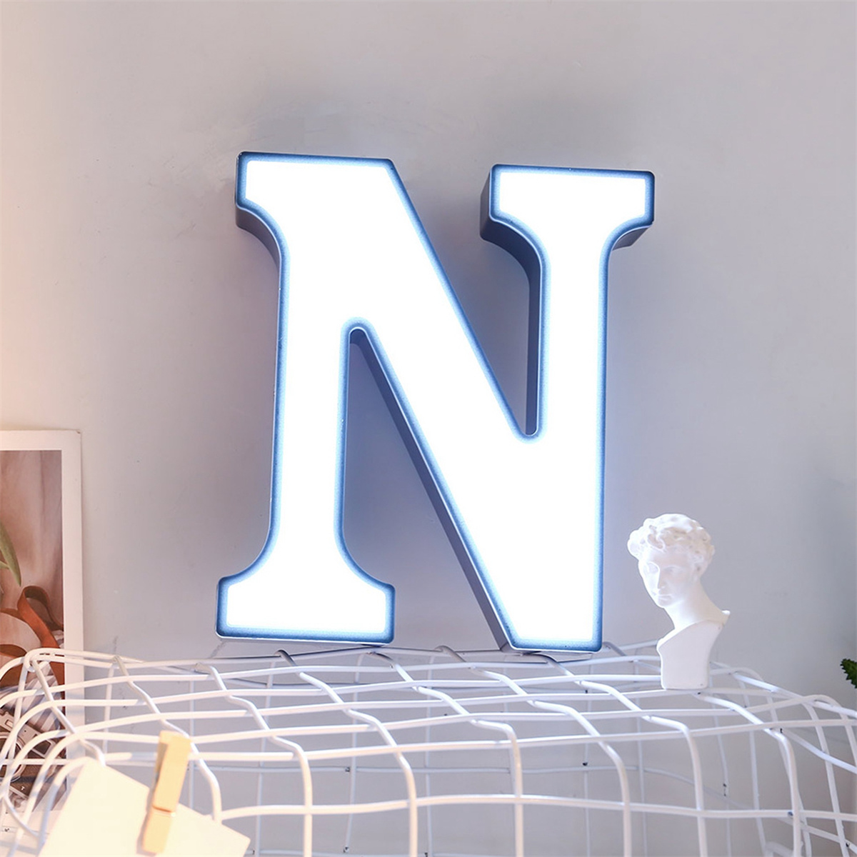 Weiß beleuchteter Buchstabe N an einer grauen Wand. Deko auf einem Regal darunter, mit einer kleinen Büste.