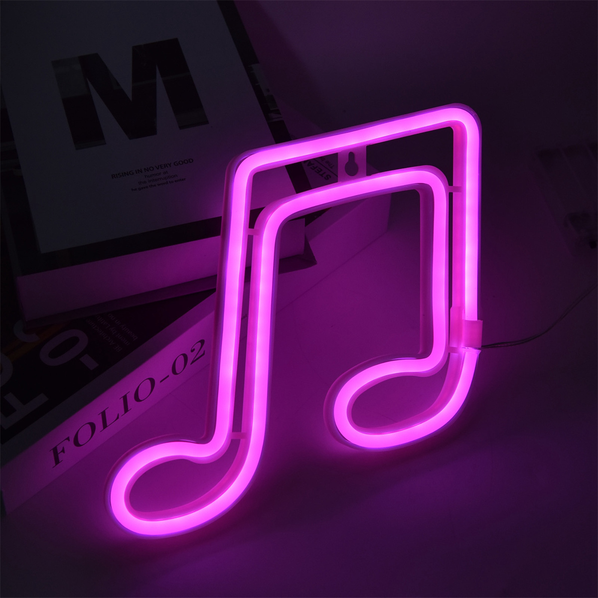 Eine neonrosa Musiknotenleuchte auf einer weißen Oberfläche. Zwei Bücher sind im Hintergrund.