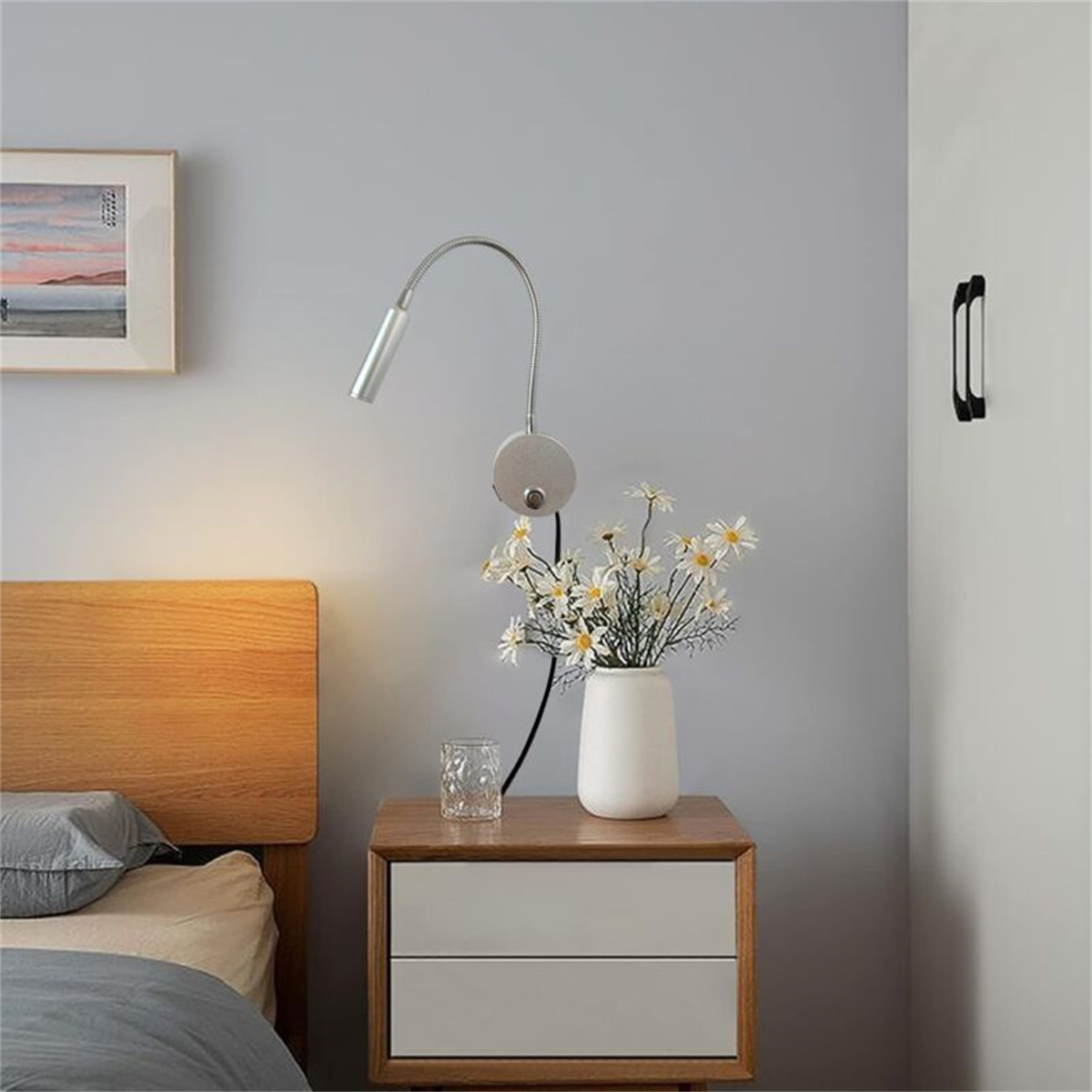 Ein Nachttisch mit Lampe, Blumenvase und einer an der Wand montierten Leselampe darüber.