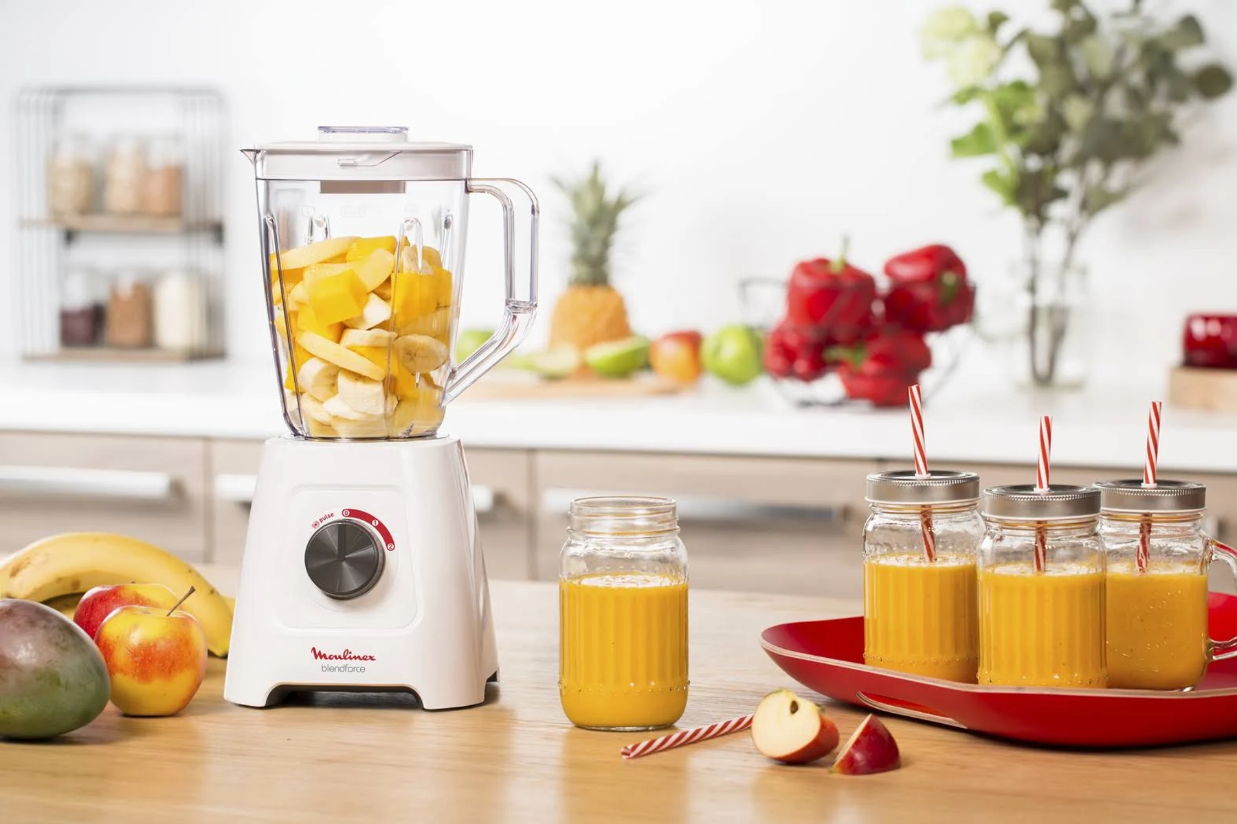 Un frullatore bianco e rosso con frutta all'interno e tre bicchieri di succo su un vassoio.