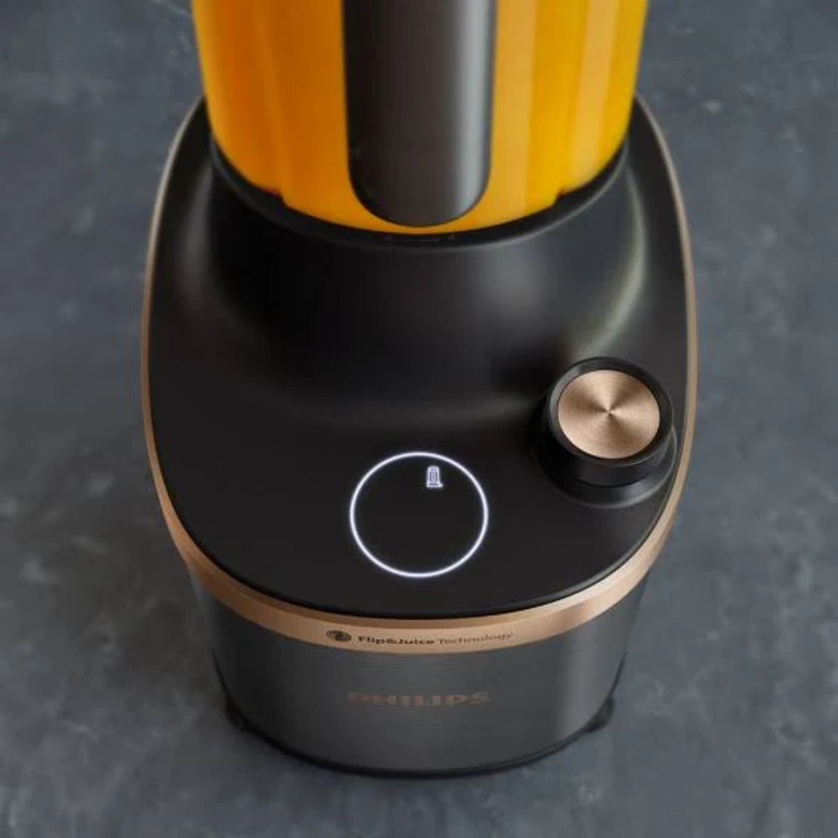 Zbliżenie czarno-złotej podstawy blendera z pokrętłem i przyciskiem. Pomarańczowy płyn w dzbanku.