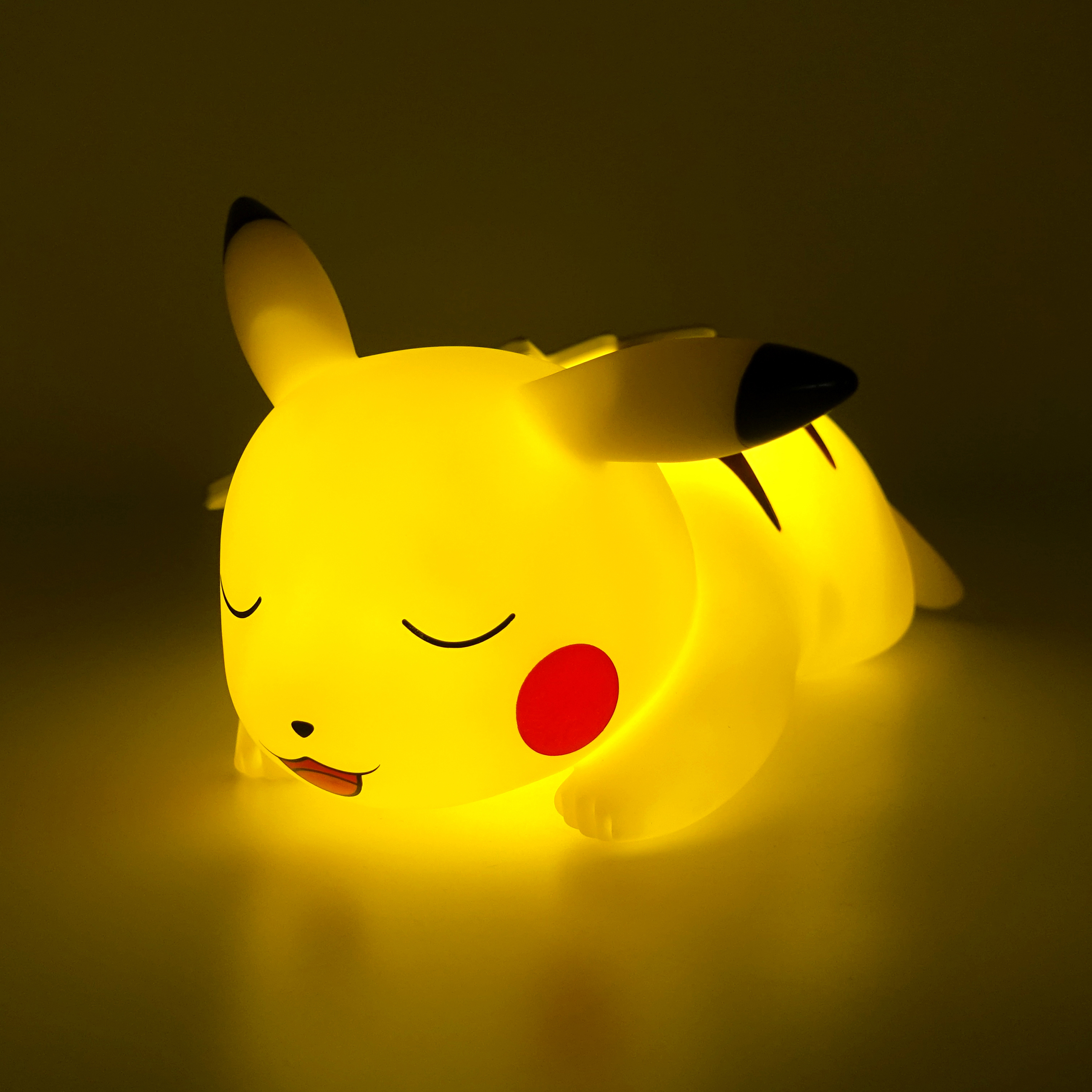 Lampada Pikachu luminosa, sdraiata con occhi chiusi e macchia rossa sulla guancia, sfondo scuro.