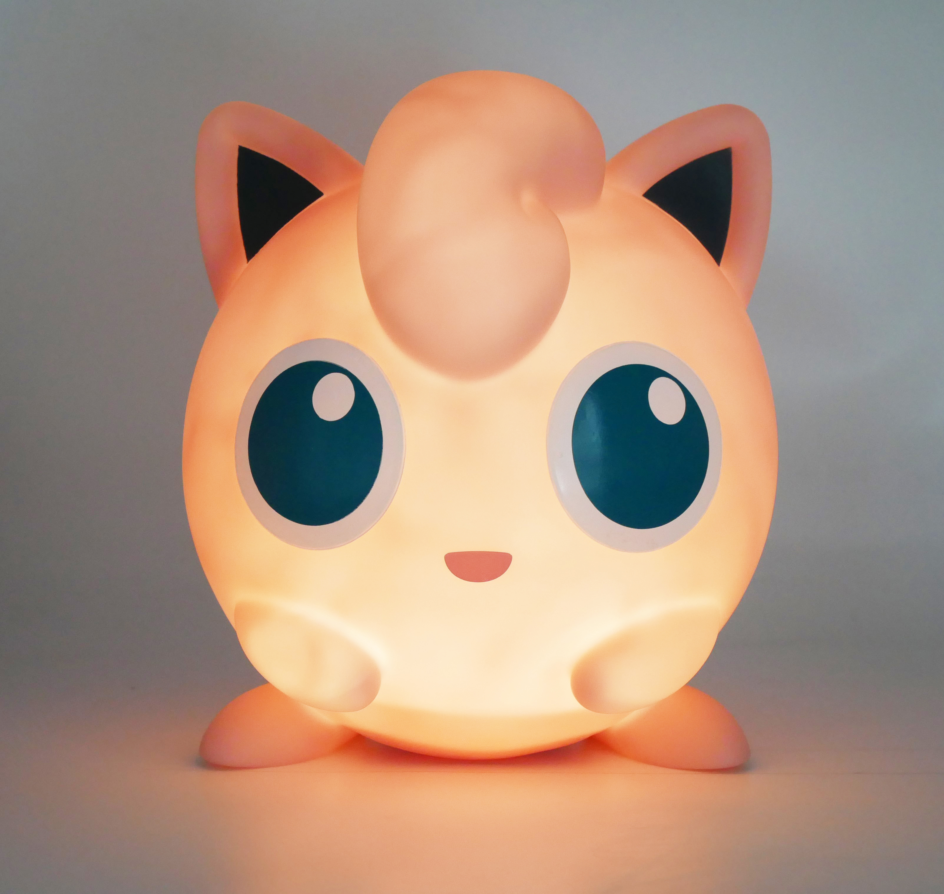 Una lampada Jigglypuff illuminata. È rotonda e rosa con orecchie nere e occhi blu.