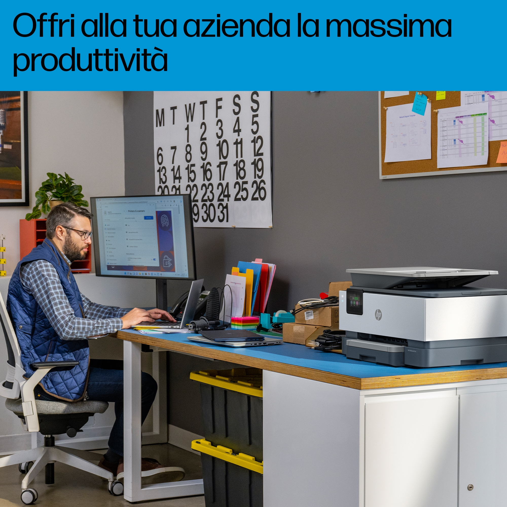 Hombre trabajando en una computadora, impresora a la derecha. Calendario y notas en la pared.