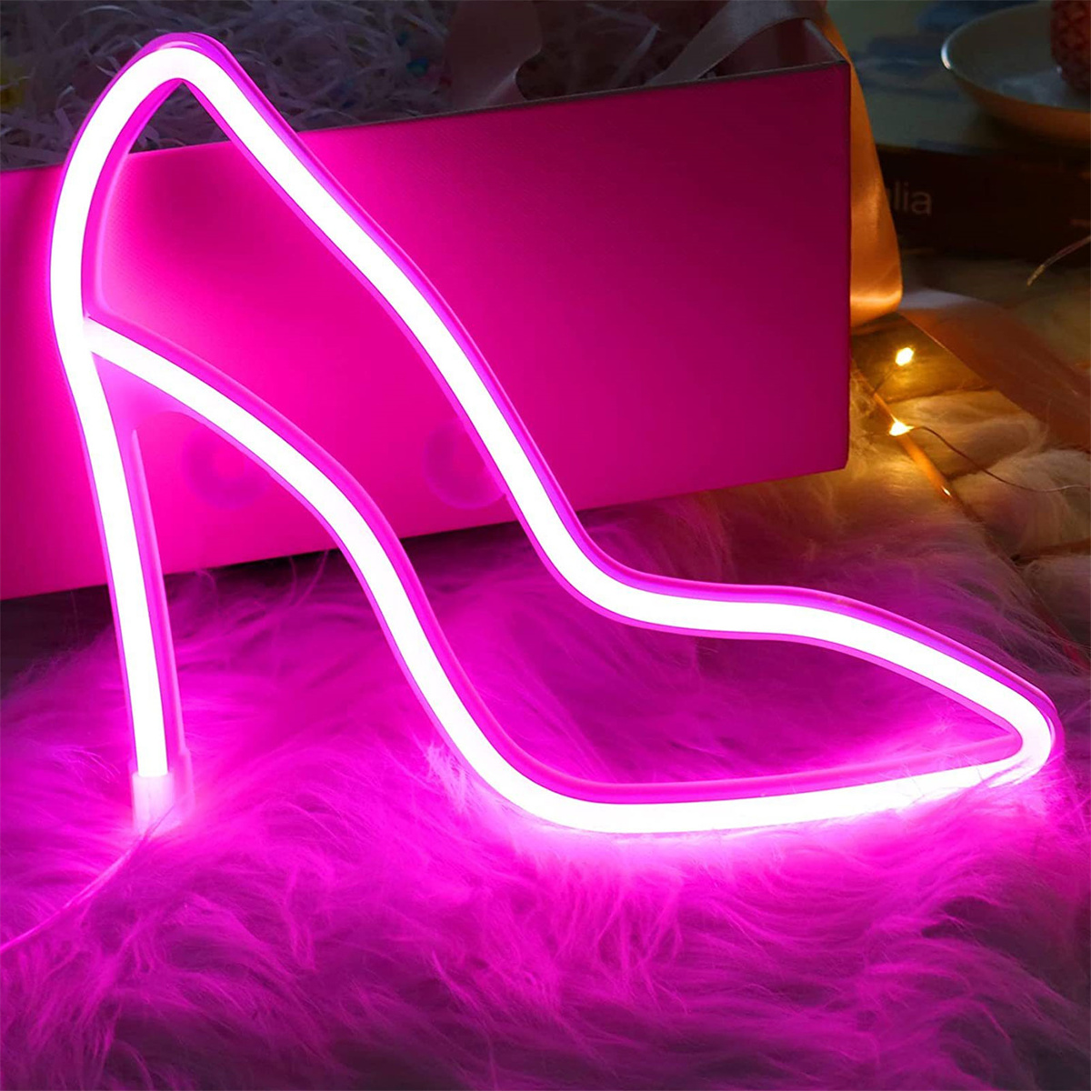 Neonpinkfarbene High-Heel-Schuh-Leuchte. Sie steht auf einer rosa Pelzoberfläche mit einer Geschenkbox im Hintergrund.