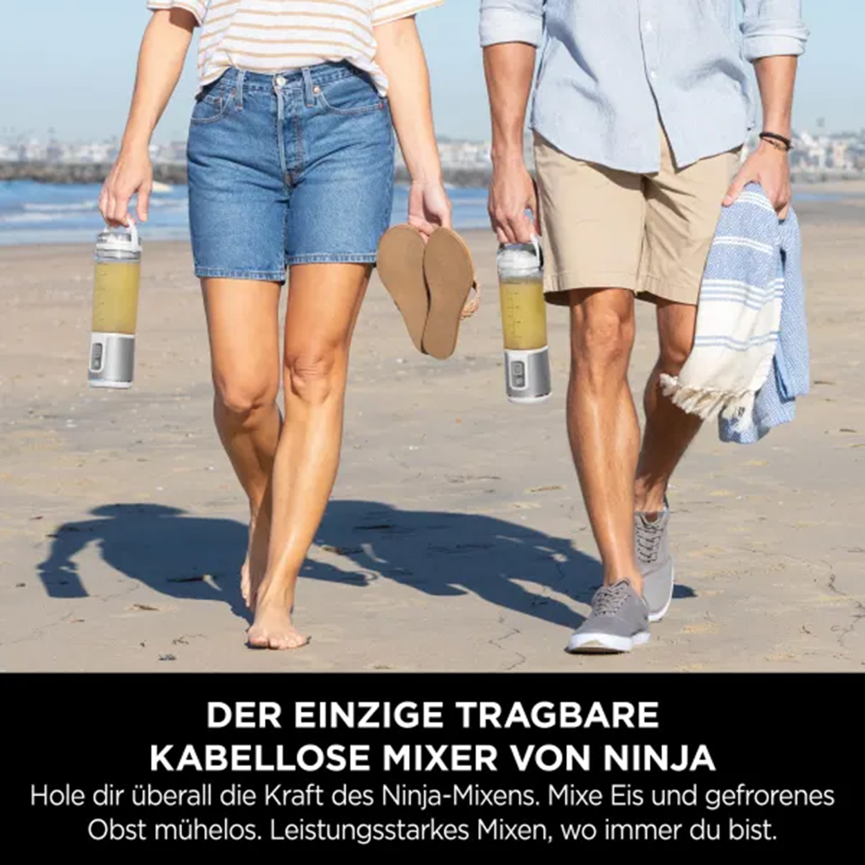 Zwei Personen gehen am Strand. Einer hält einen Mixer, der andere ein Handtuch und Sandalen.