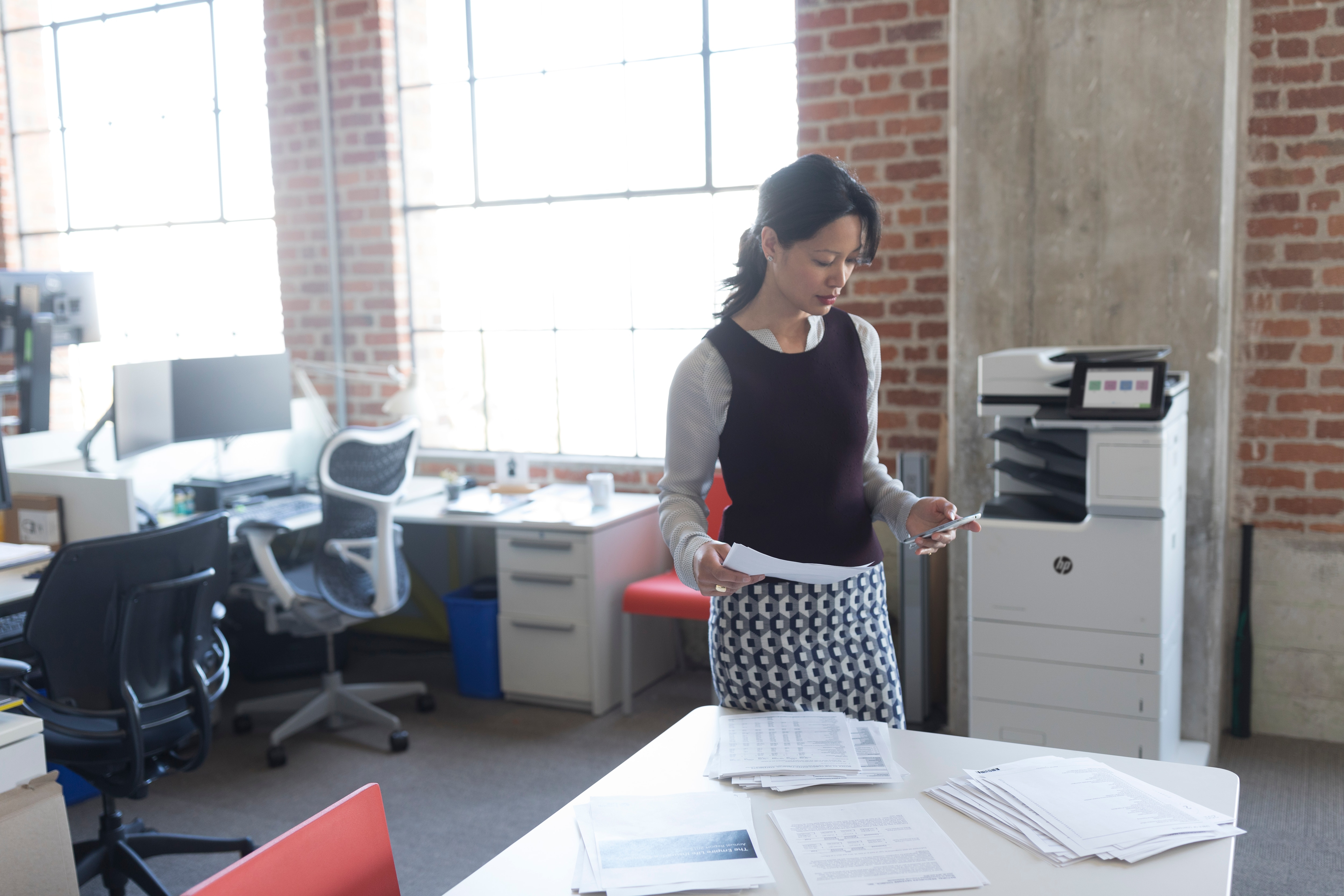 Kobieta w biznesowym stroju stoi przy drukarce w biurze. Trzyma papiery.