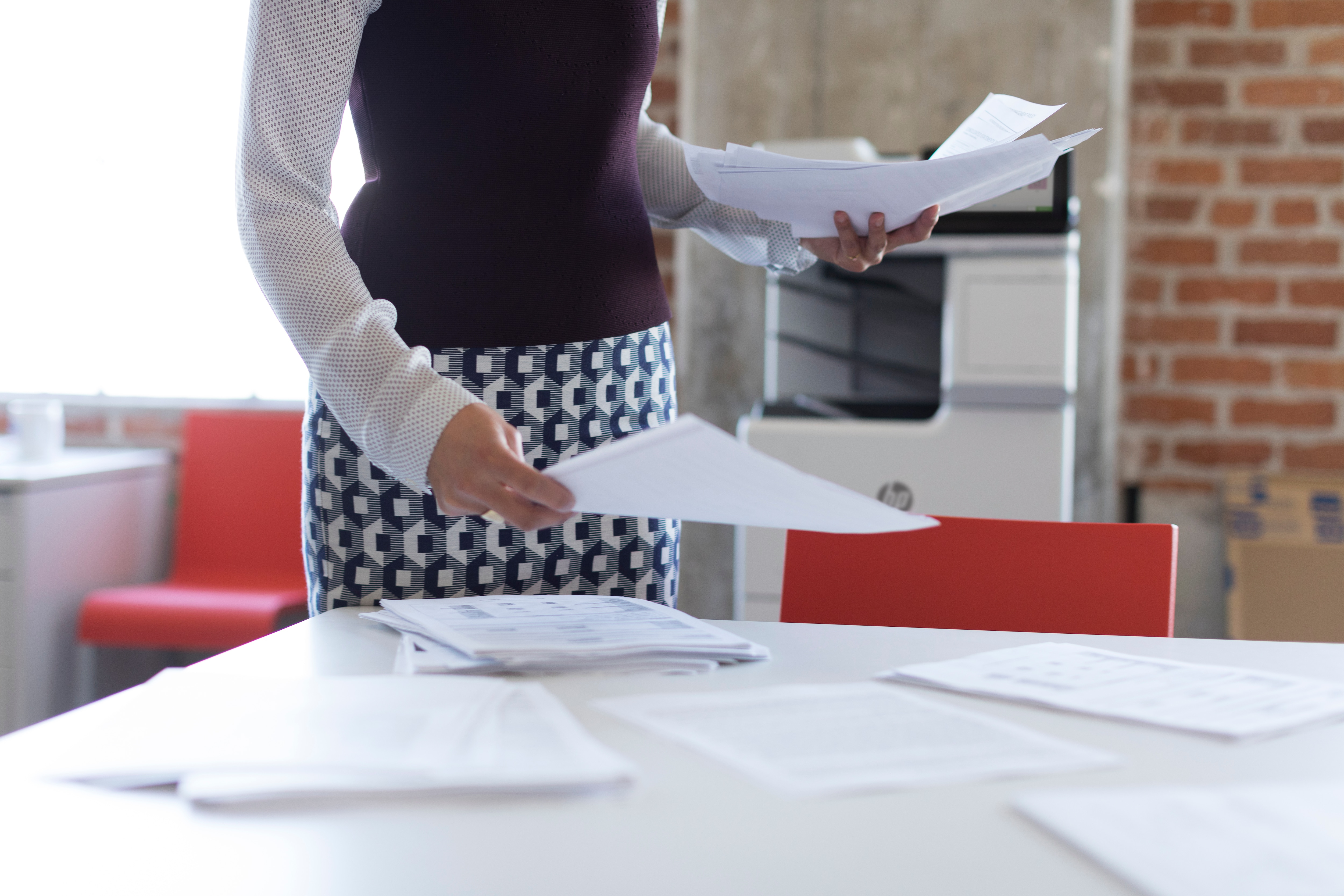 Zbliżenie kobiety trzymającej papiery. Drukarka i czerwone krzesło w tle.