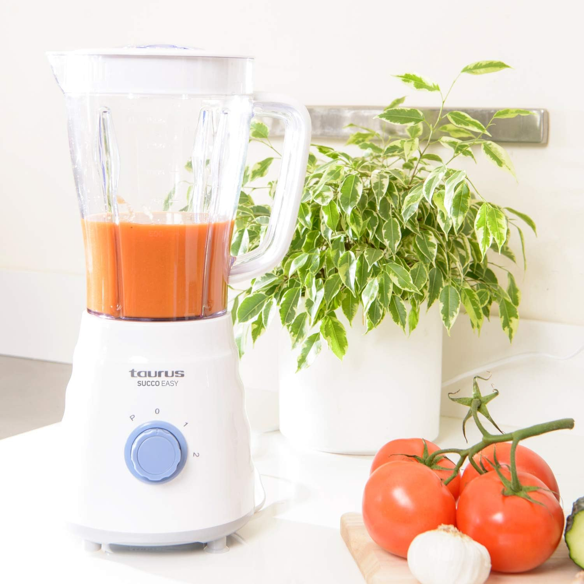 Licuadora con líquido naranja junto a tomates y una planta.