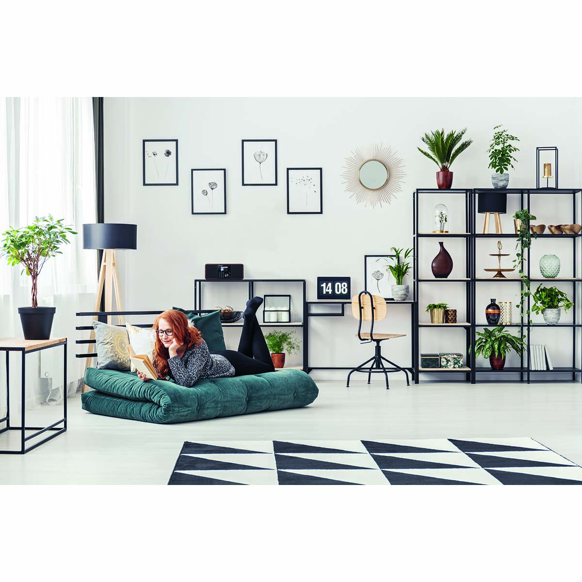 A woman on a teal futon reading a book. There are plants and decorations around her.