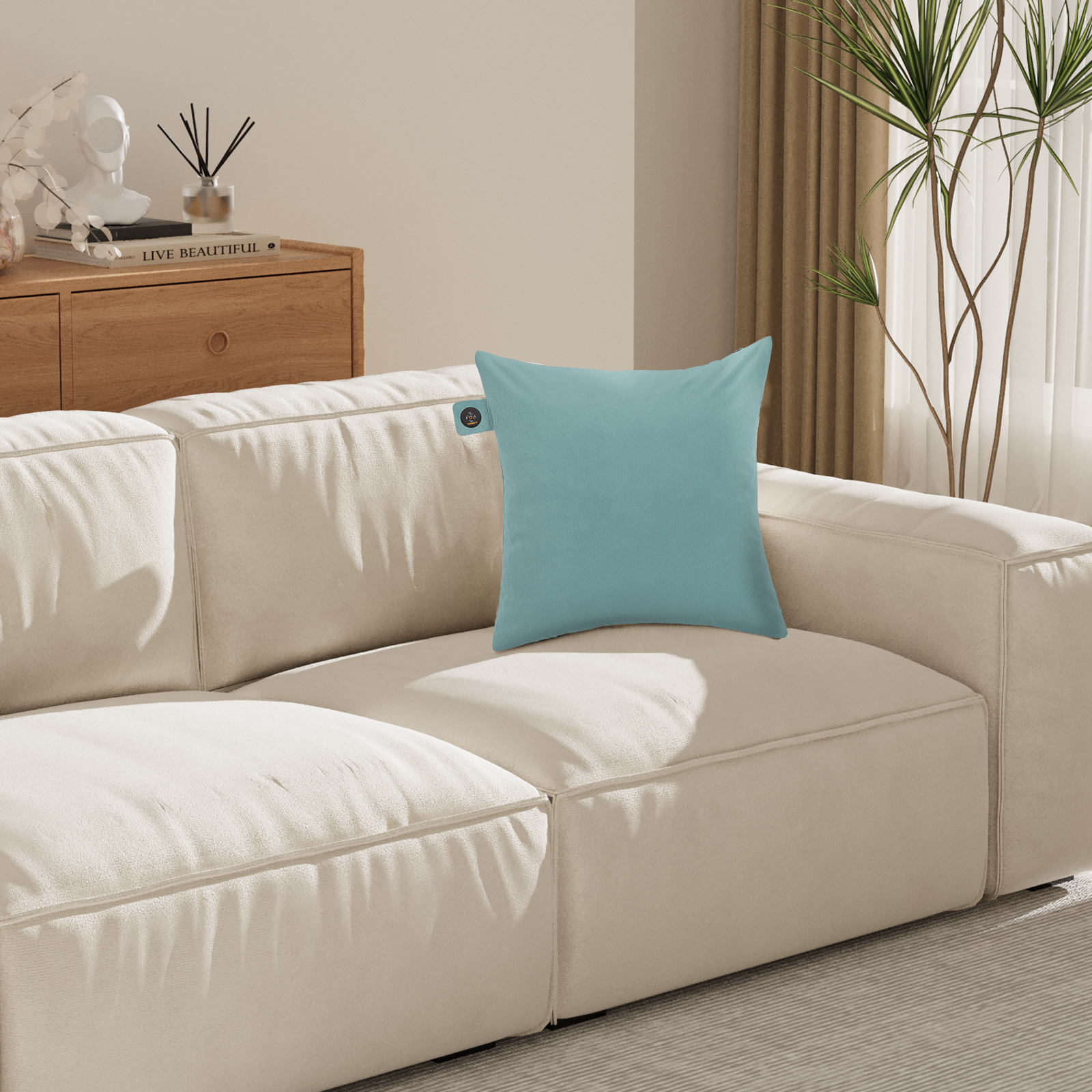 Ein blaues quadratisches Kissen auf einem cremefarbenen Sofa. Ein Holzschrank und eine Pflanze befinden sich im Hintergrund.