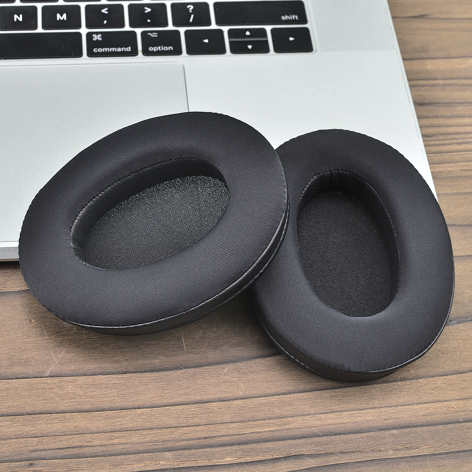 Dwie czarne owalne poduszki do słuchawek na drewnianej powierzchni, obok klawiatury laptopa.