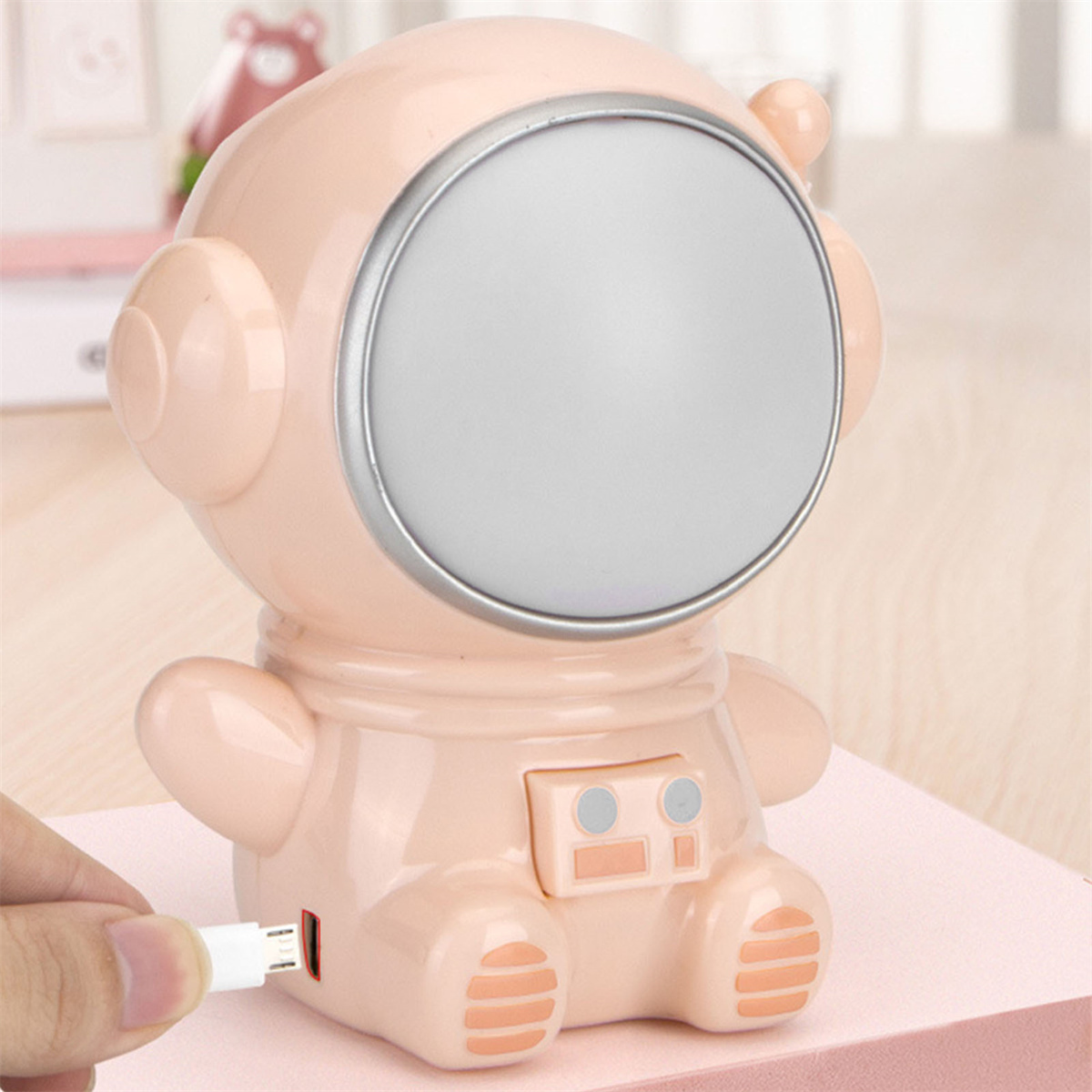 Rosa astronautenförmige Lampe mit großem, rundem Licht. Eine Hand steckt ein USB-Kabel ein.