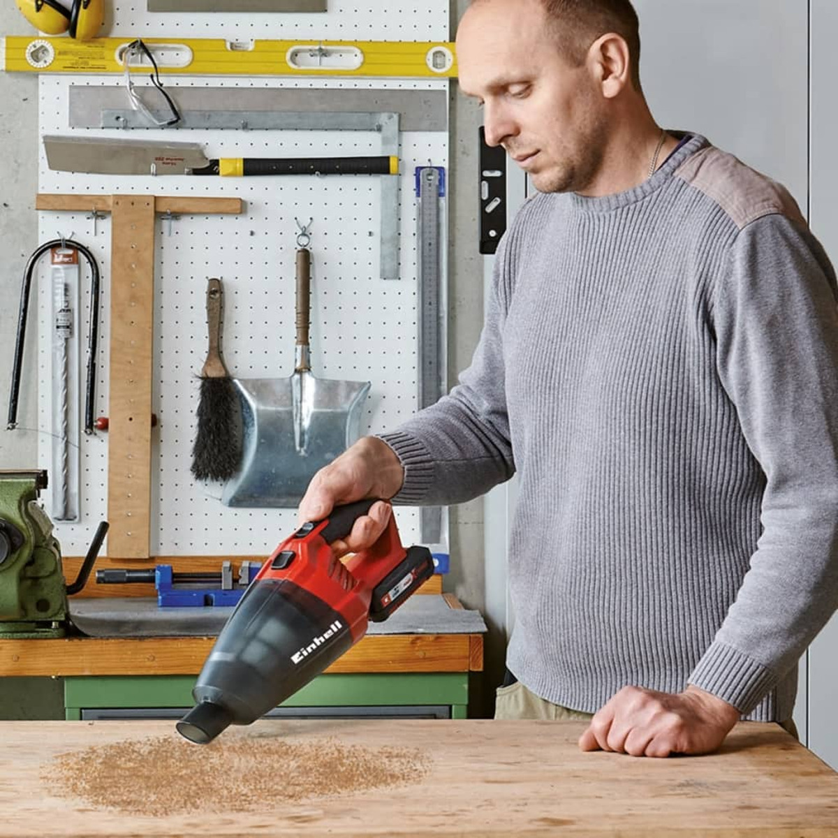 Mężczyzna używa czerwonego odkurzacza ręcznego na drewnianym stole warsztatowym, obecny pył.