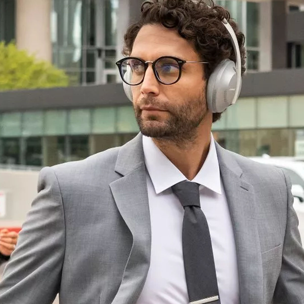 Ein Mann im grauen Anzug mit Brille und Kopfhörern, der draußen in einer Stadt geht.