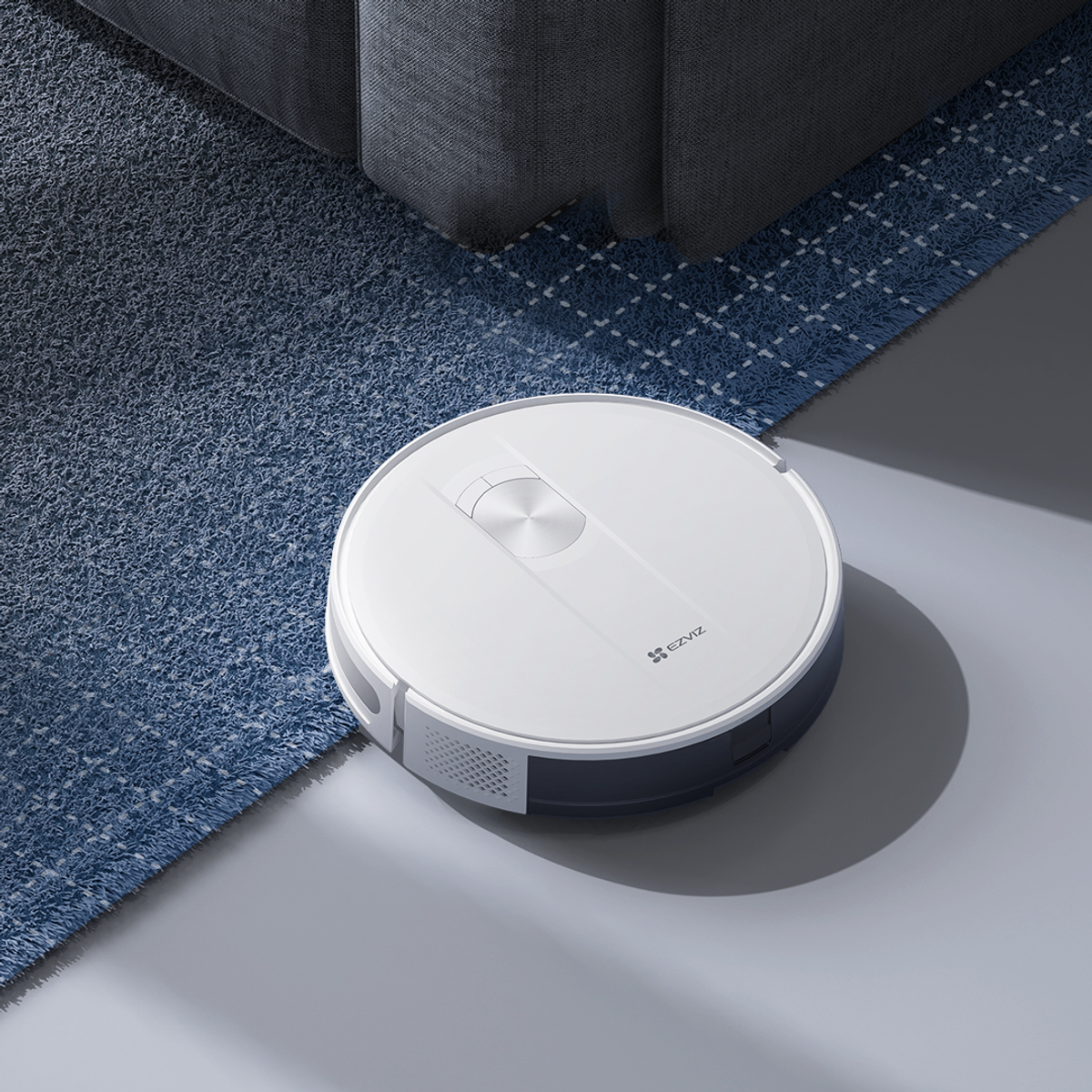 Weißer Roboter-Staubsauger auf blau gemustertem Teppich, in der Nähe eines grauen Sofas. Der Boden ist grau.