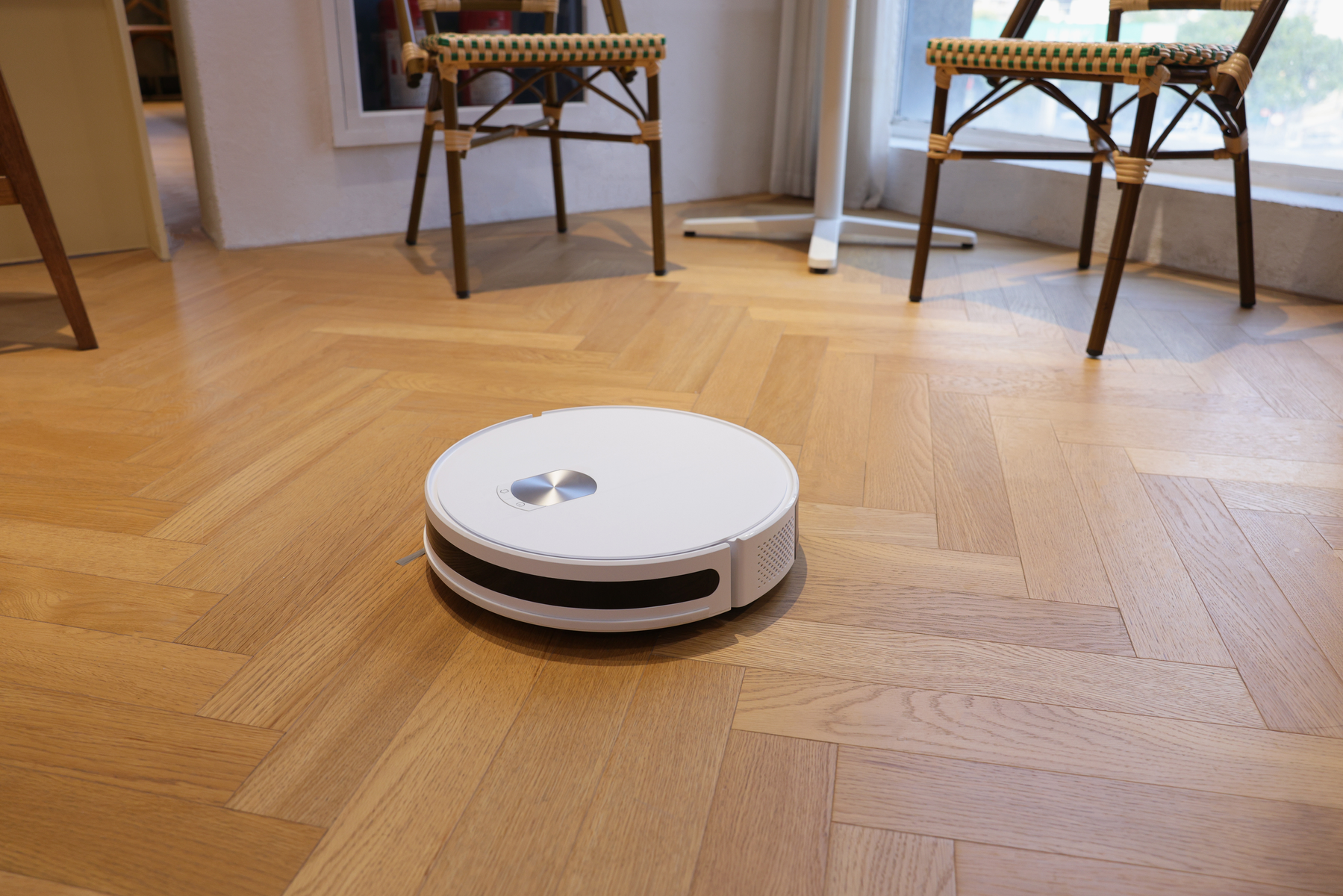 Ein weißer Roboter-Staubsauger auf einem Holzboden. Stühle im Hintergrund.