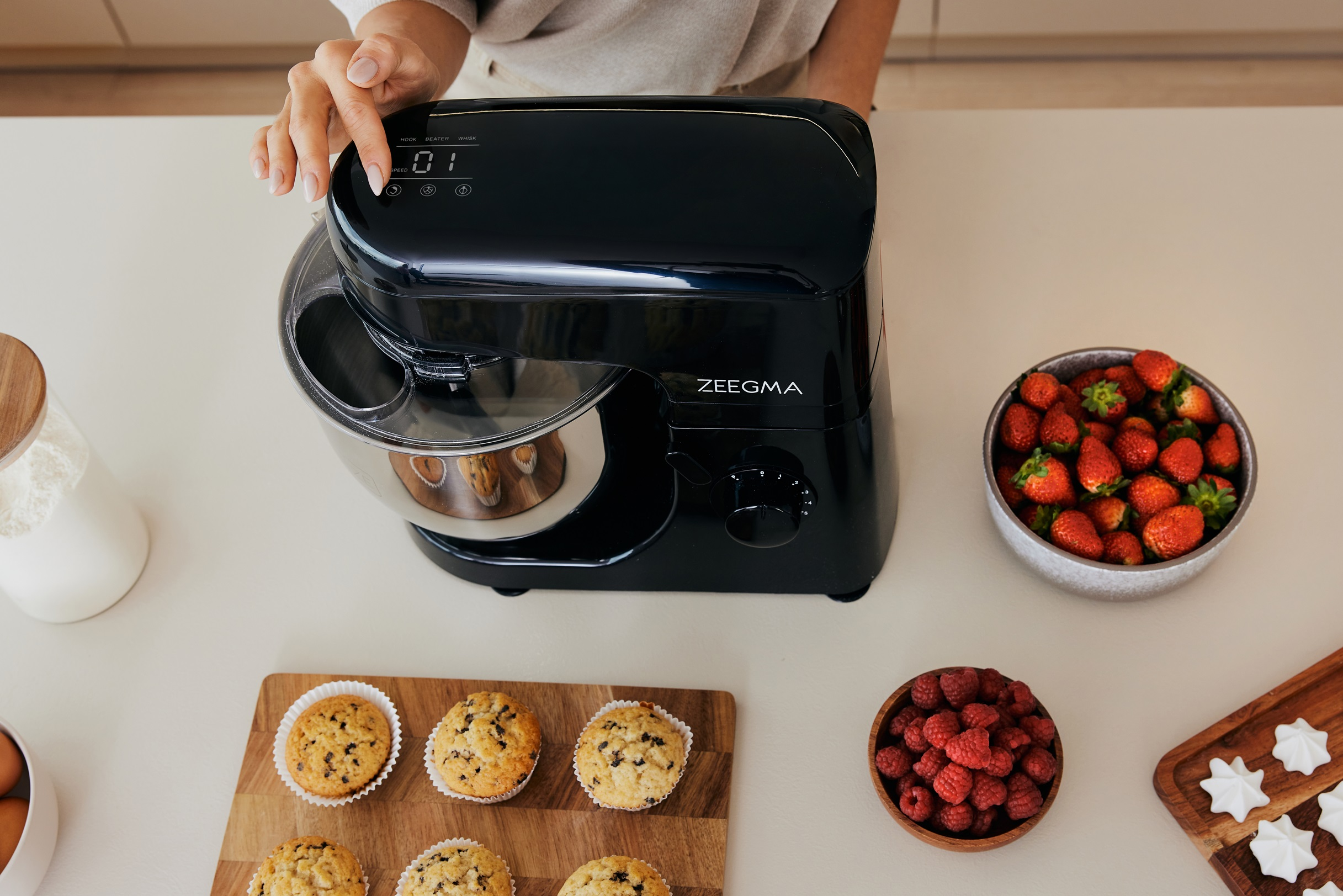 Kobieta używa czarnego miksera stojącego na białym blacie. Na blacie są jagody i babeczki.