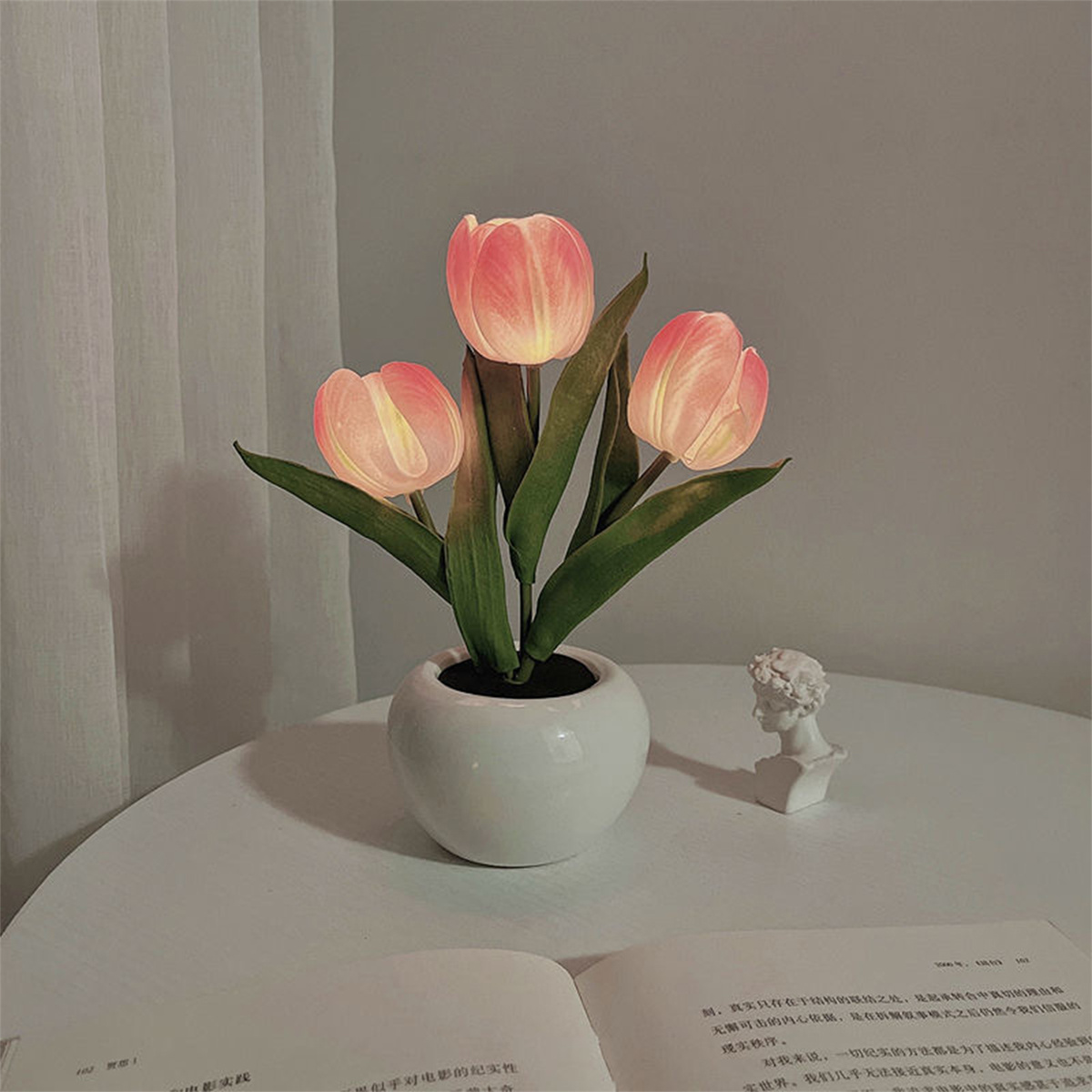 Künstliche Tulpen in einem weißen Topf auf einem Tisch mit einer kleinen Statue und einem Buch.