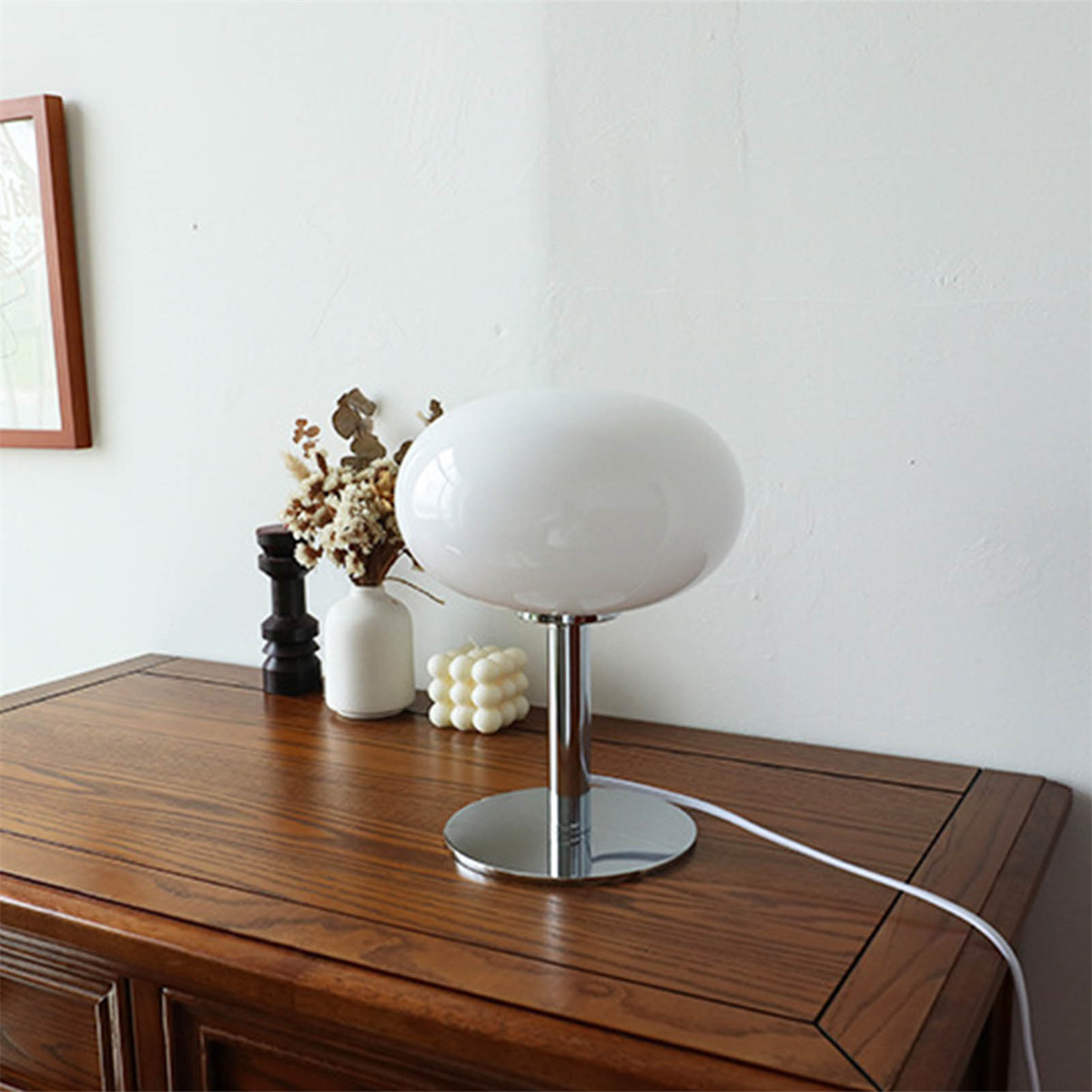 Weiße ovale Lampe auf einem dunkelbraunen Tisch. Eine Vase mit Blumen steht daneben.