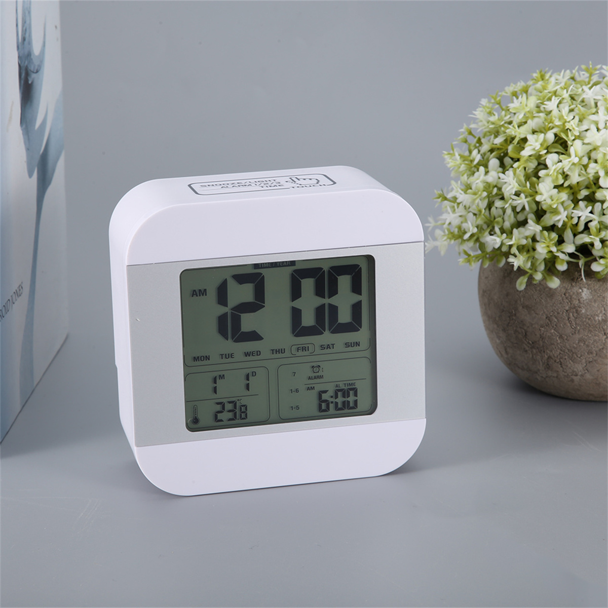 Weiße Digitaluhr mit Zeit- und Datumsanzeige, mit einer Pflanze.