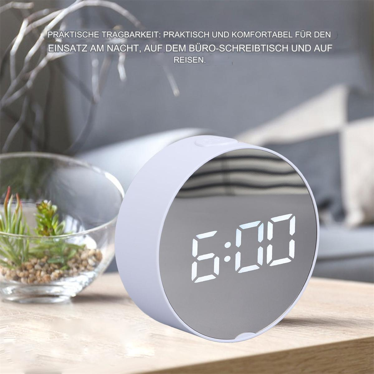 Wecker mit Digitalanzeige, die 6:00 Uhr anzeigt. Es ist ein kreisförmiges Design mit verspiegelter Oberfläche. Holztisch und Pflanzen.