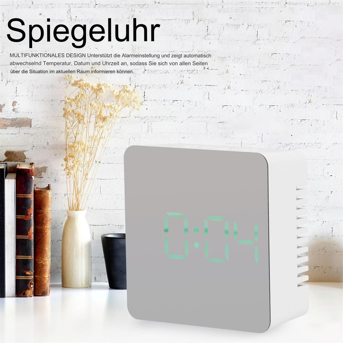 Eine verspiegelte Uhr auf einem weißen Tisch, mit einer Backsteinmauer im Hintergrund und einer Vase mit Trockenblumen.
