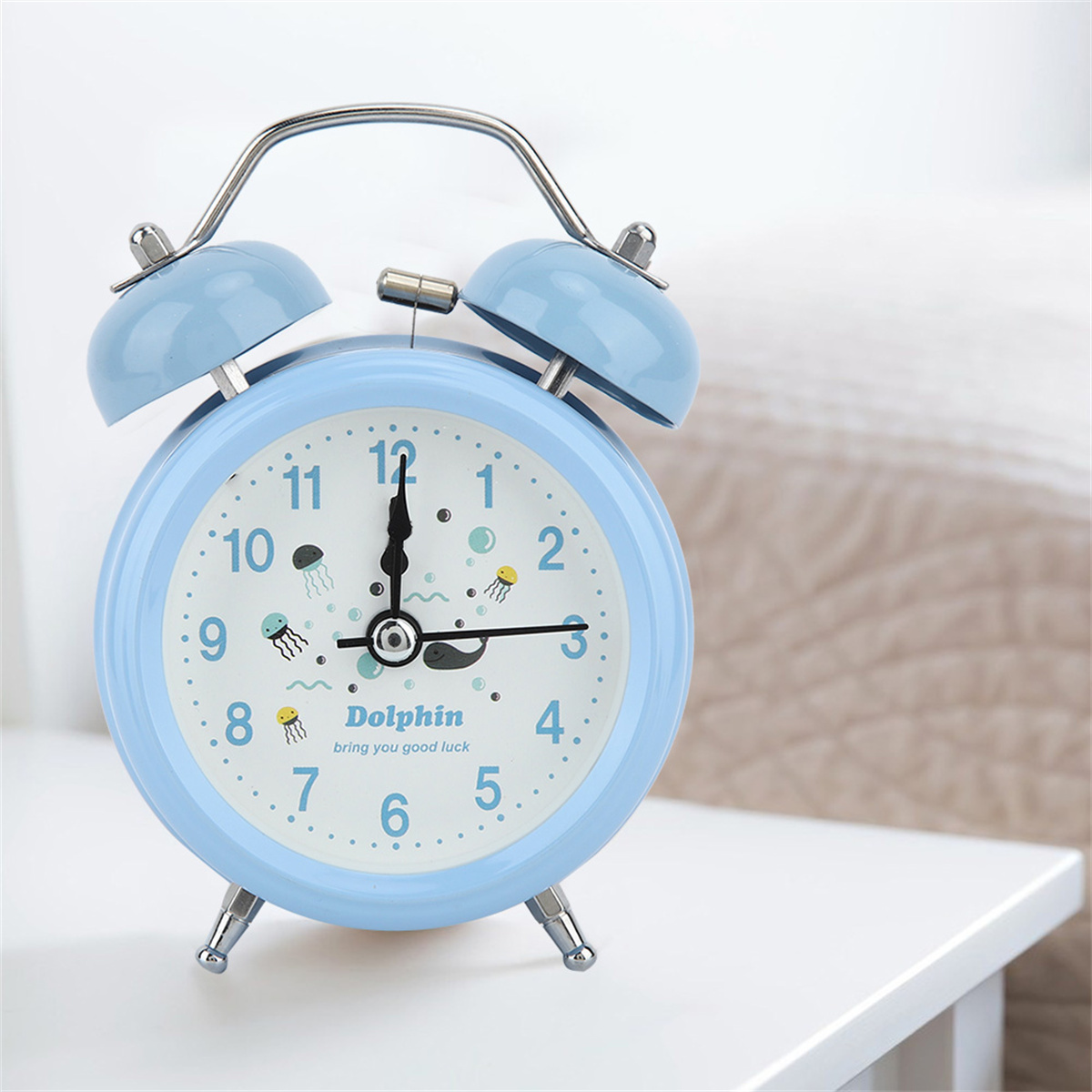 Ein blauer Wecker mit weißem Zifferblatt auf weißer Oberfläche, mit Bett im Hintergrund.
