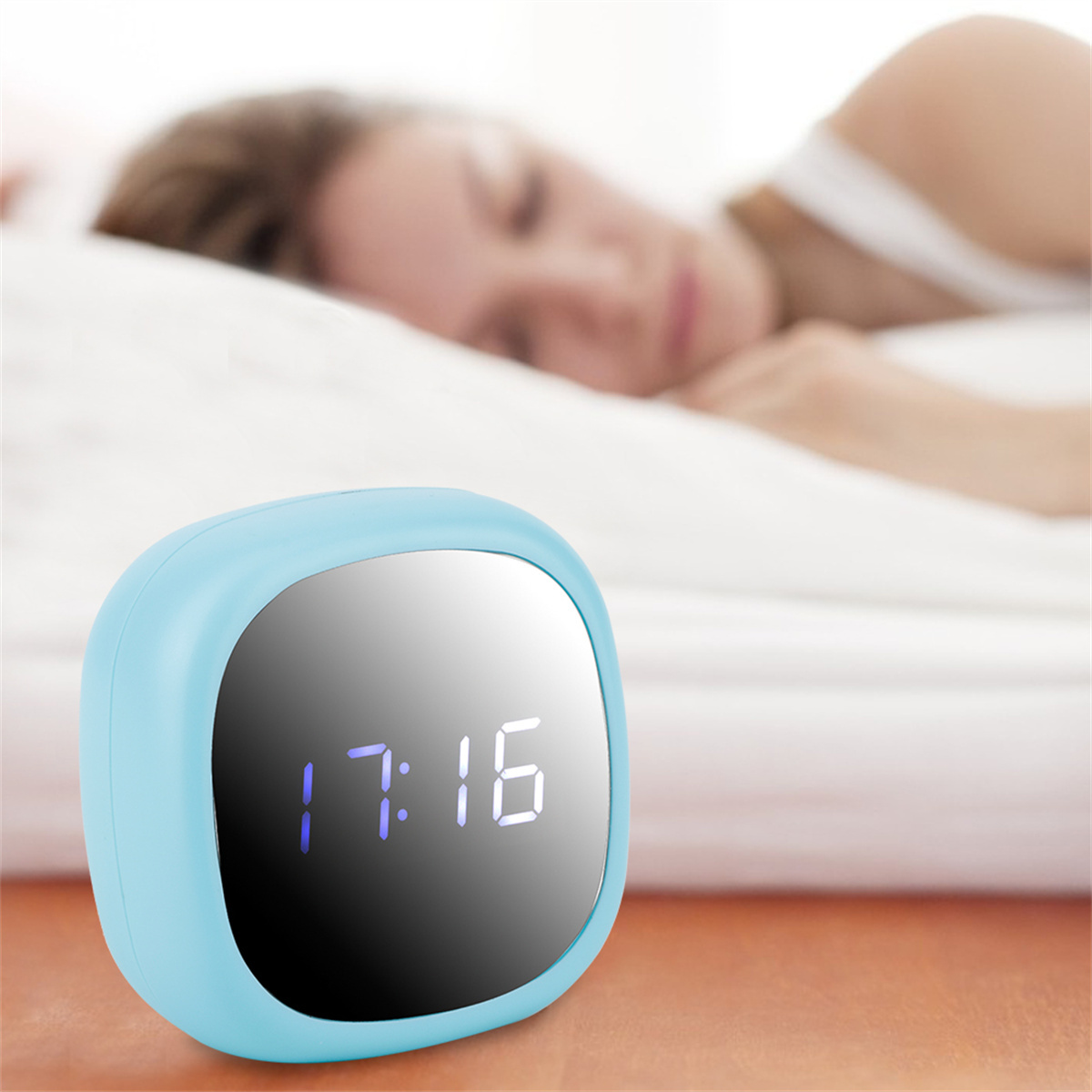 Ein hellblauer Wecker zeigt 17:16, neben einer schlafenden Person in einem weißen Bett.