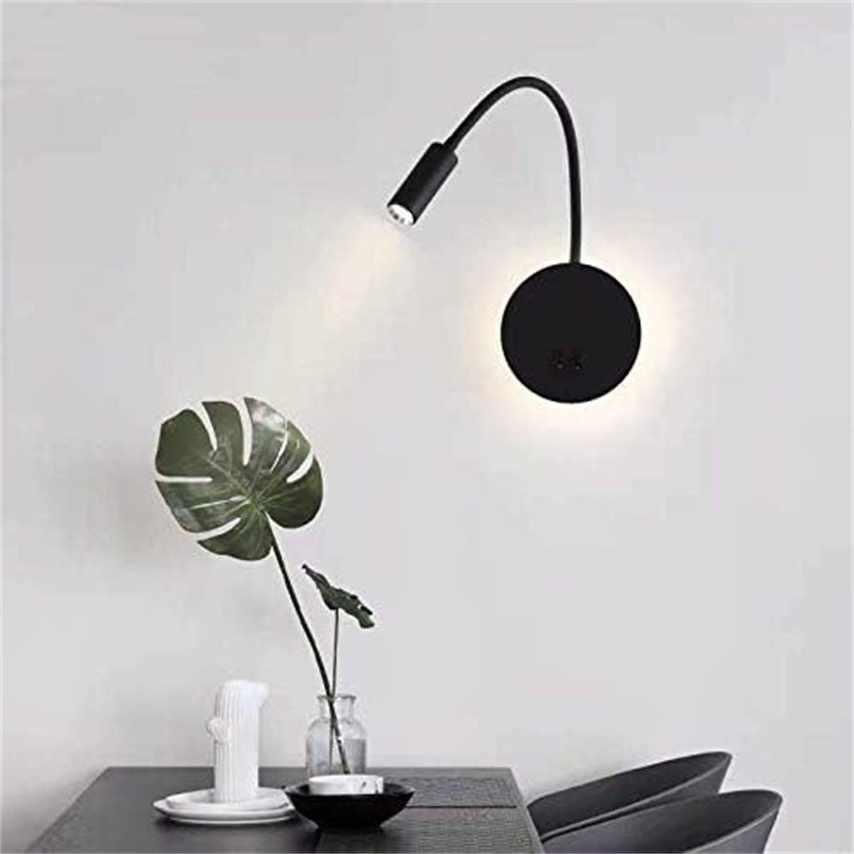 Eine schwarze Wandlampe mit gebogenem Arm. Daneben eine grüne Blätterpflanze.