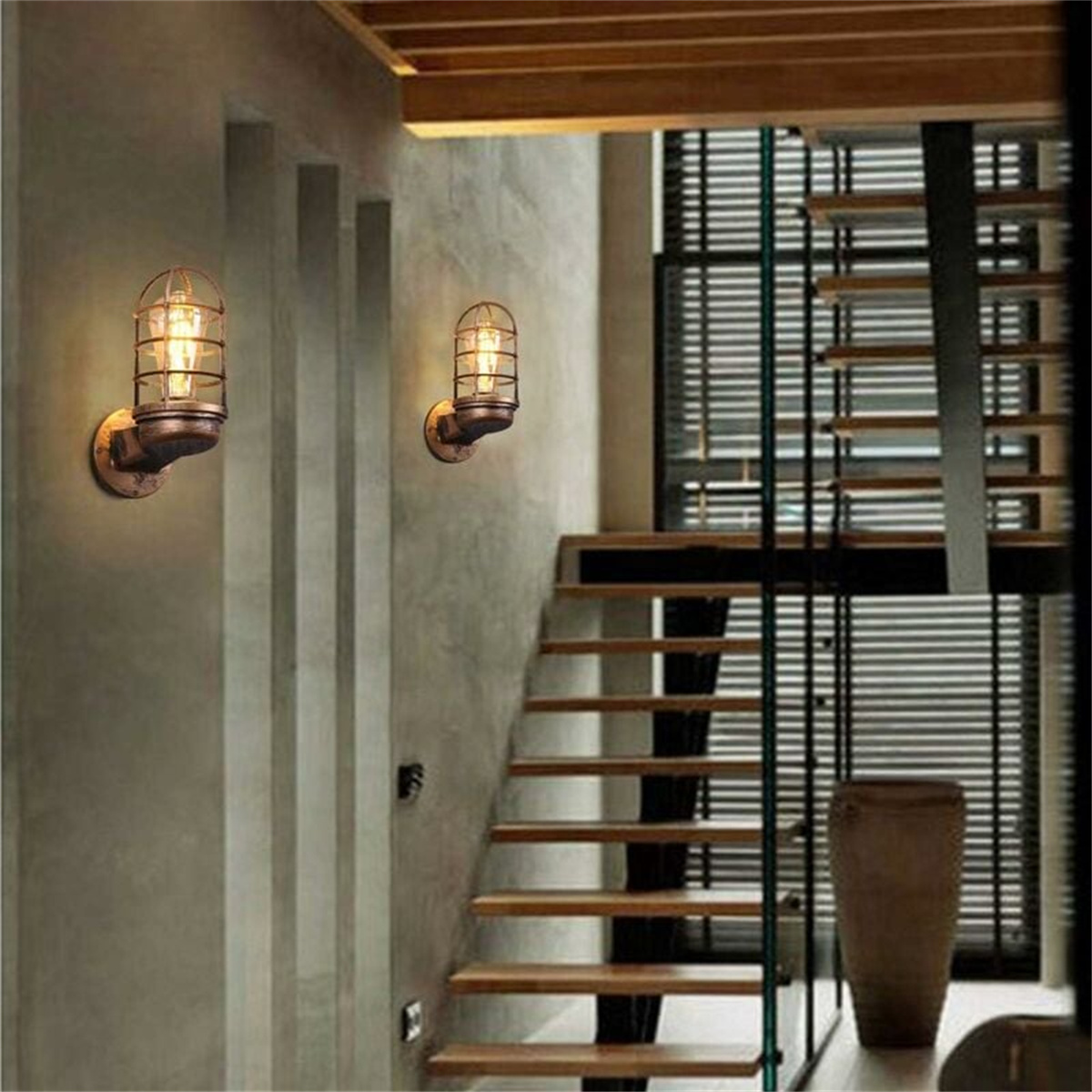 Zwei Wandleuchten mit Käfigbirnen. Treppe mit Glaspaneelen und eine hohe braune Vase.