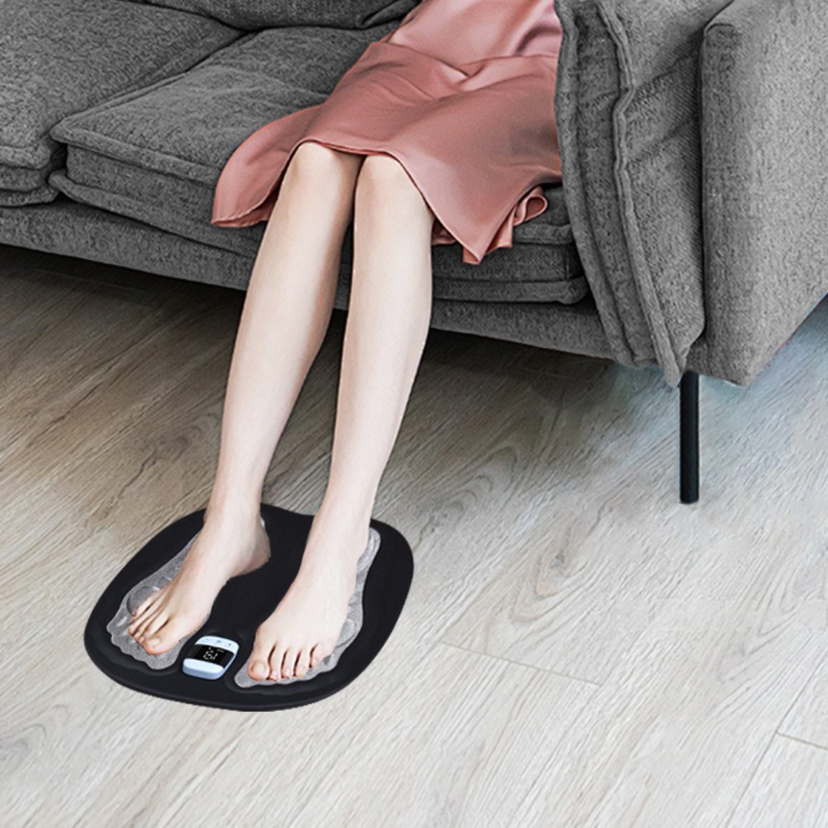 Beine einer Person, Füße auf einem Fußmassagegerät, sitzend auf einem grauen Sofa, heller Holzboden.