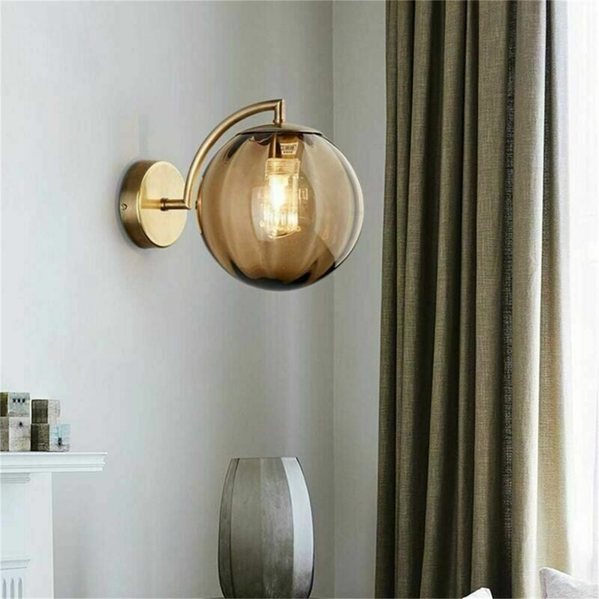 Eine Wandleuchte mit goldenem Sockel und einem runden, bernsteinfarbenen Glasschirm. Die Wand ist hellgrau mit einem grünen Vorhang.