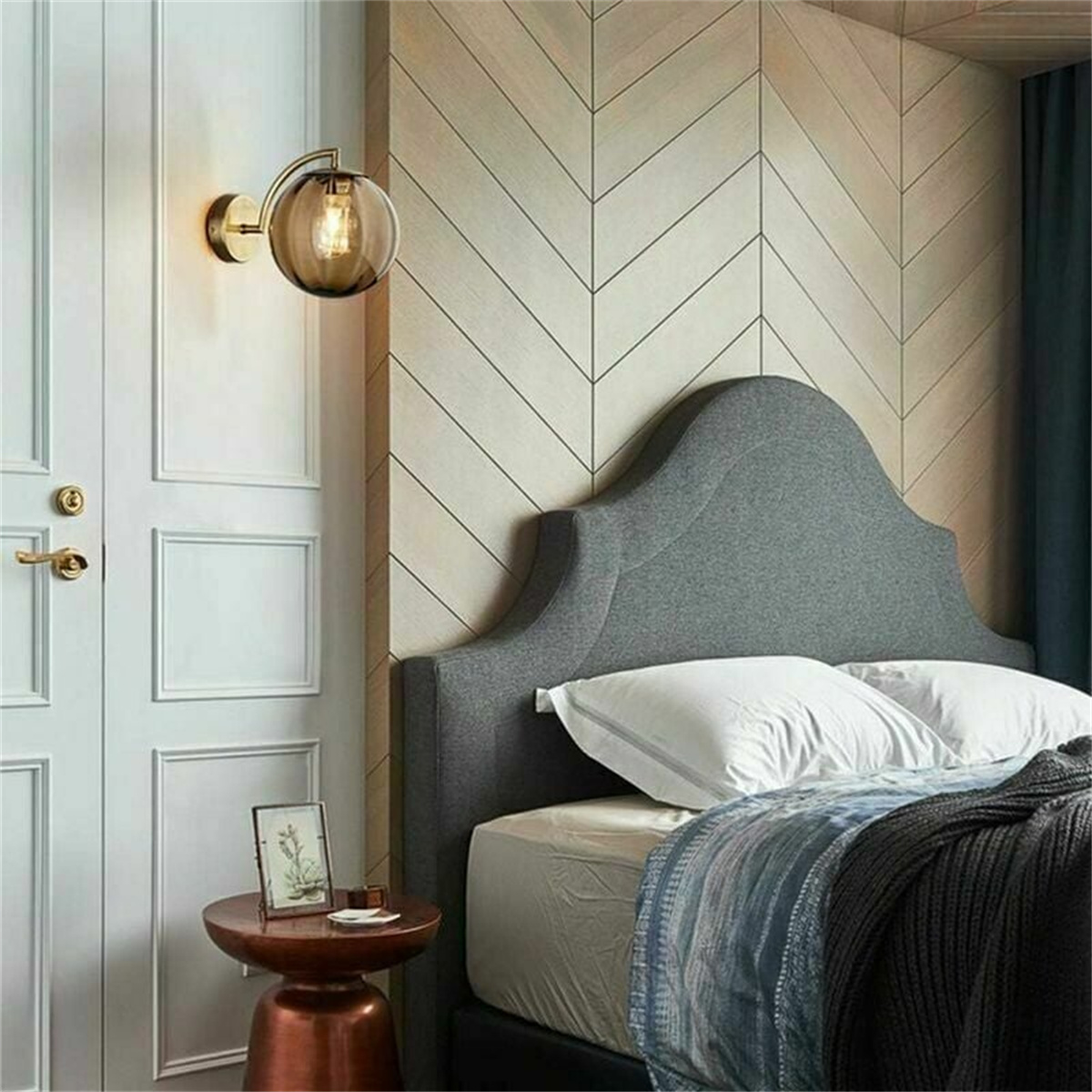 Eine Wandleuchte und ein Bett mit Kissen und einem grauen Kopfteil. Die Wand hat ein Fischgrätmuster.