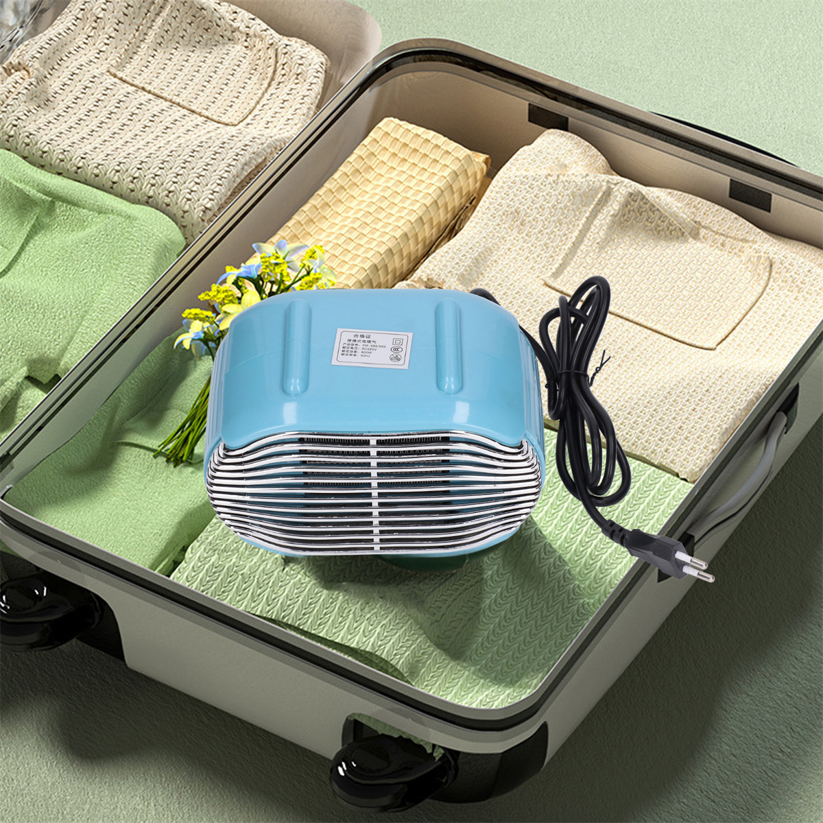 Ein blauer kleiner Heizlüfter in einem offenen Koffer. Kleidung ist darum gepackt. Mit schwarzem Kabel.