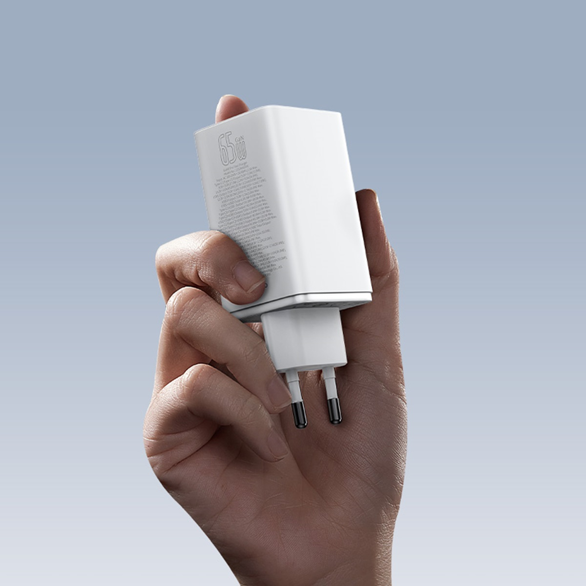 Une main tient un chargeur blanc avec le texte '65W'. Fond de ciel bleu.