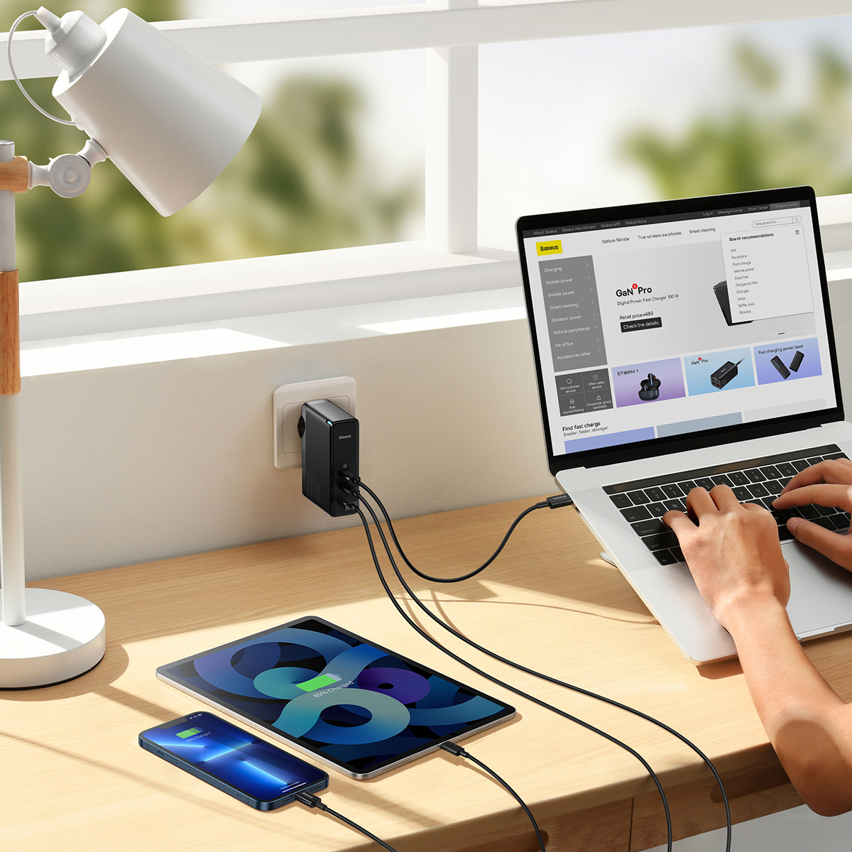 Biurko z laptopem, tabletem i telefonem w trakcie ładowania. Widoczna jest również lampa i zasilacz.