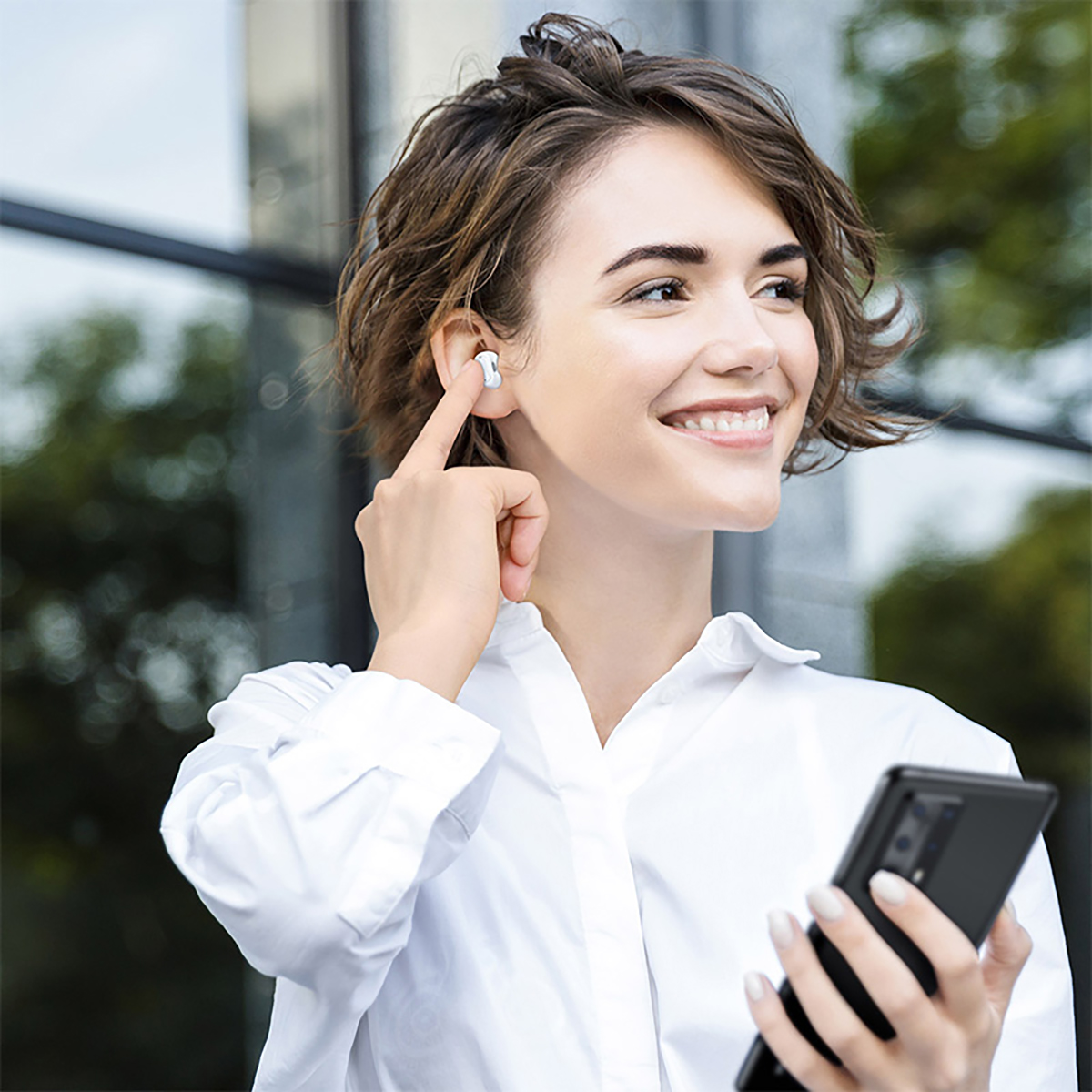 Donna con auricolari, tiene in mano un telefono. Sorride ed è fuori, alberi sullo sfondo.