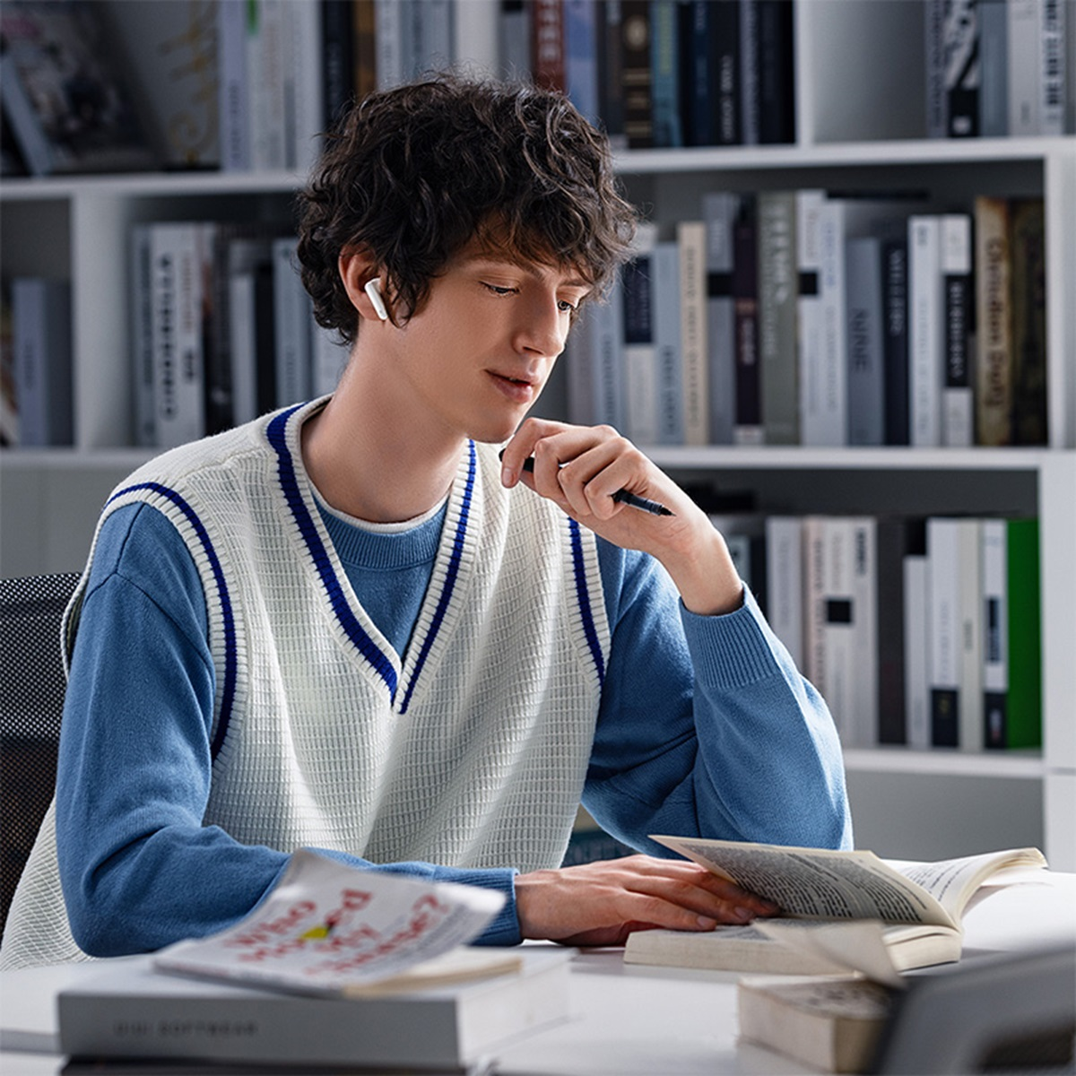 Mężczyzna w białych słuchawkach, czyta książkę. Siedzi w pokoju z regałem na książki.