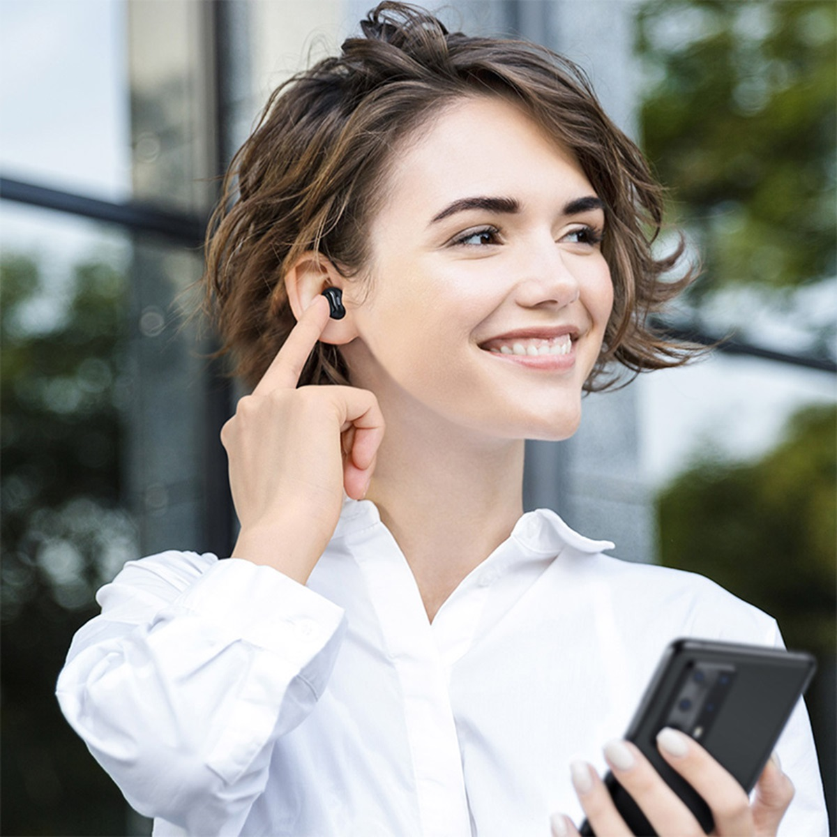 Kobieta z czarną słuchawką w uchu, trzymająca telefon, uśmiechnięta na zewnątrz.