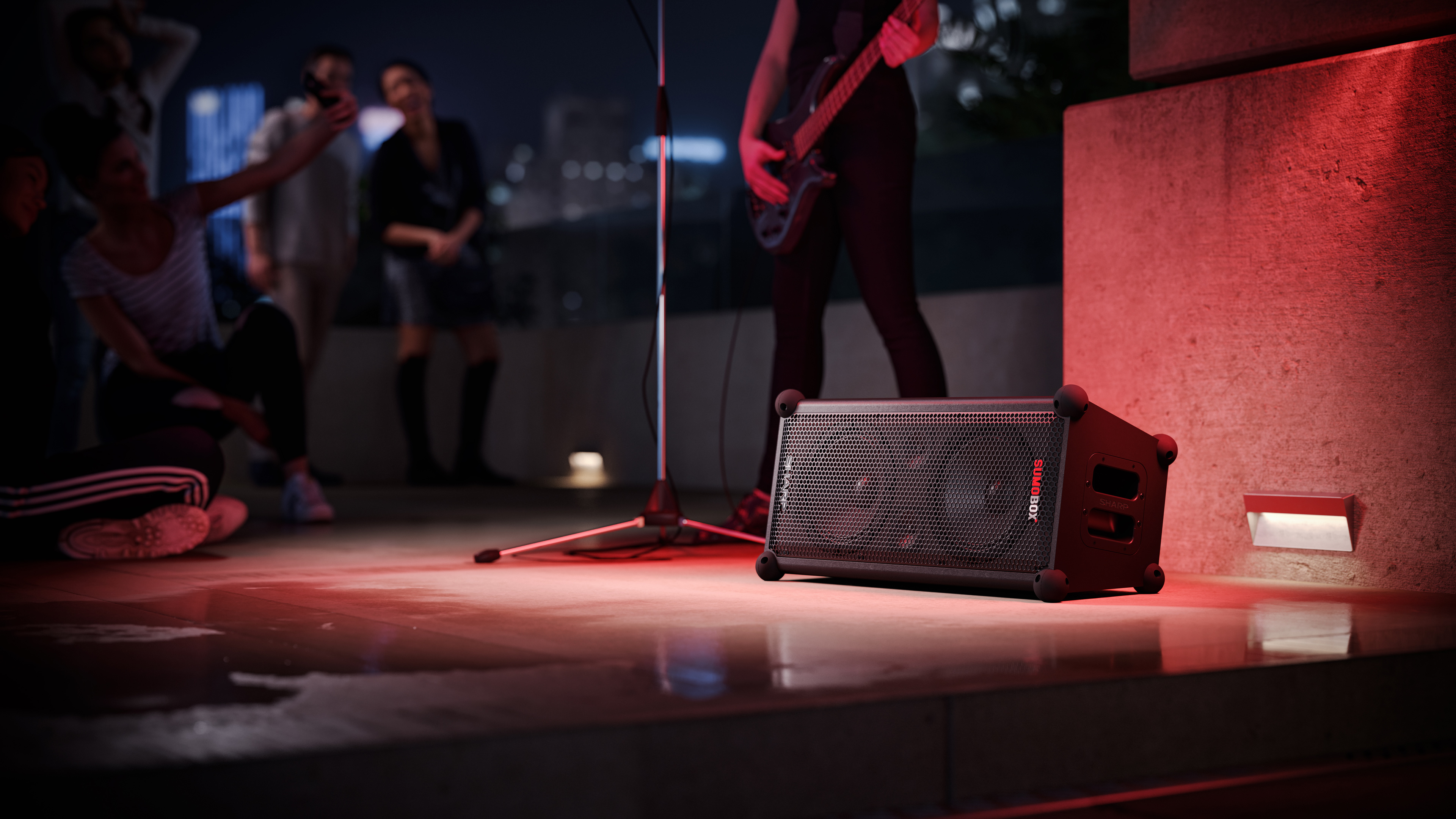 Głośnik na scenie z ludźmi w tle. Czerwone oświetlenie sceniczne. Statyw na gitarę i mikrofon.