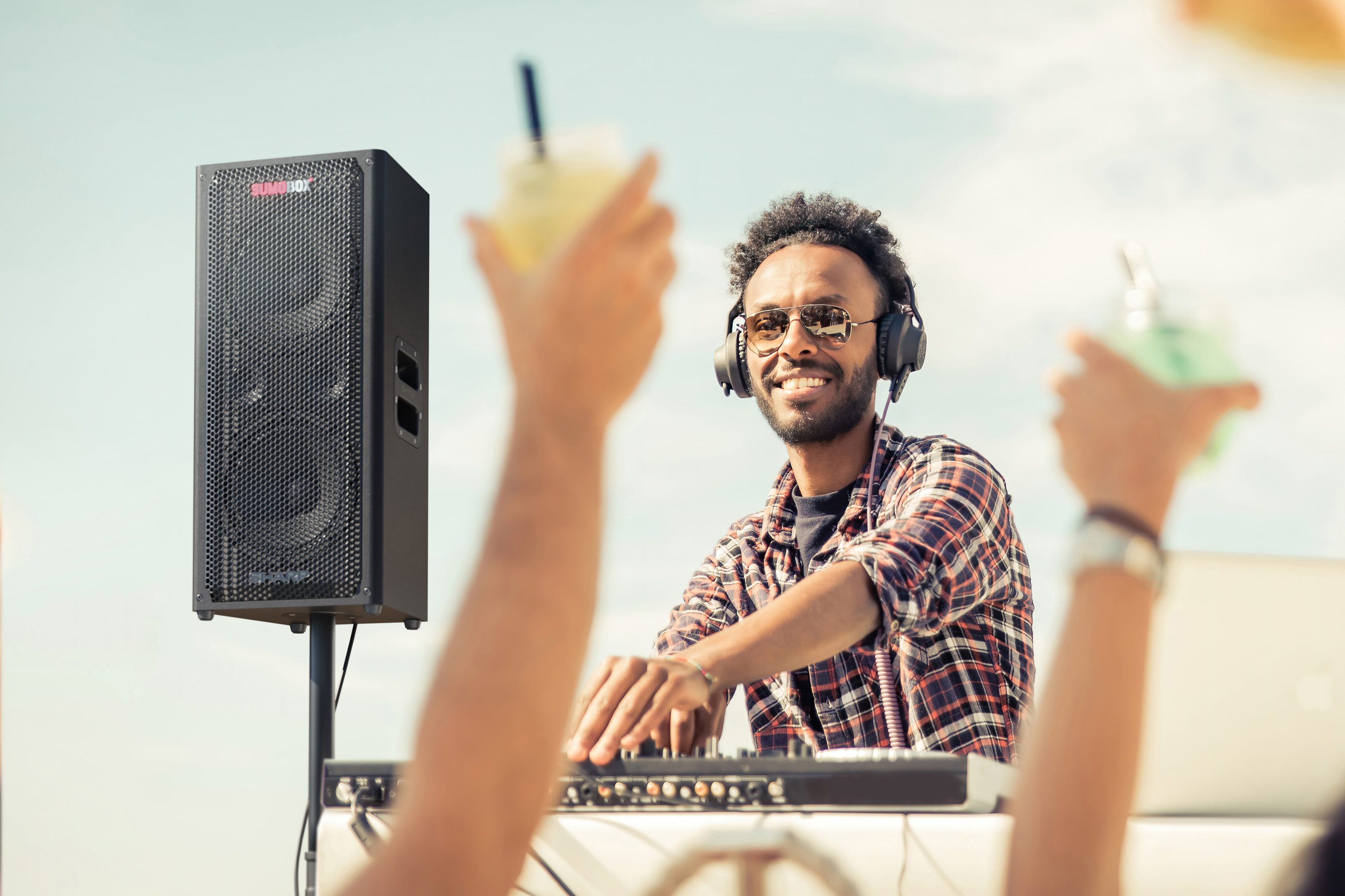 DJ na imprezie, uśmiechnięty i trzymający drinka. Głośnik i ludzie w tle.