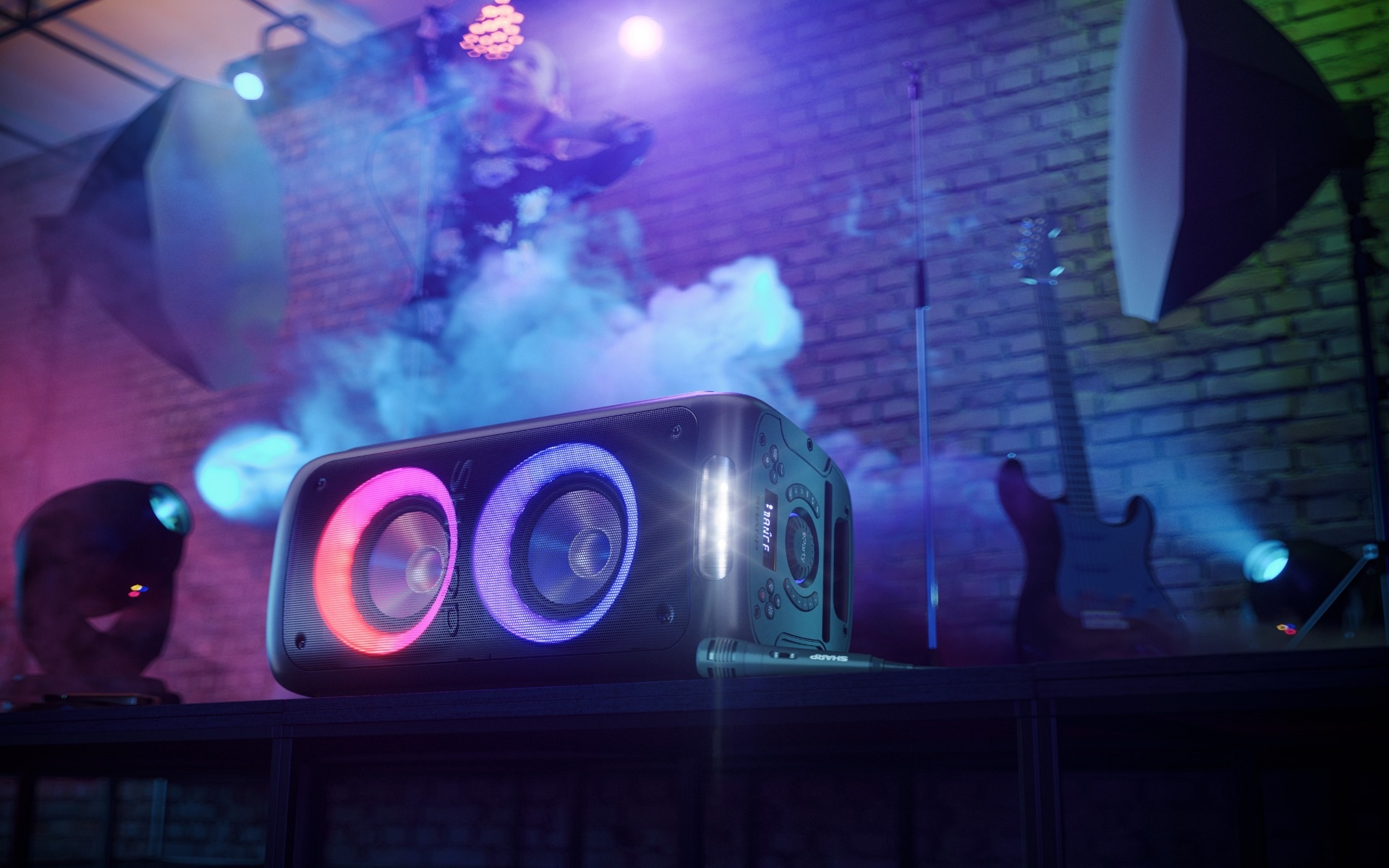 Czarny głośnik z dwoma pierścieniami świetlnymi, mikrofon na statywie i gitara w tle.